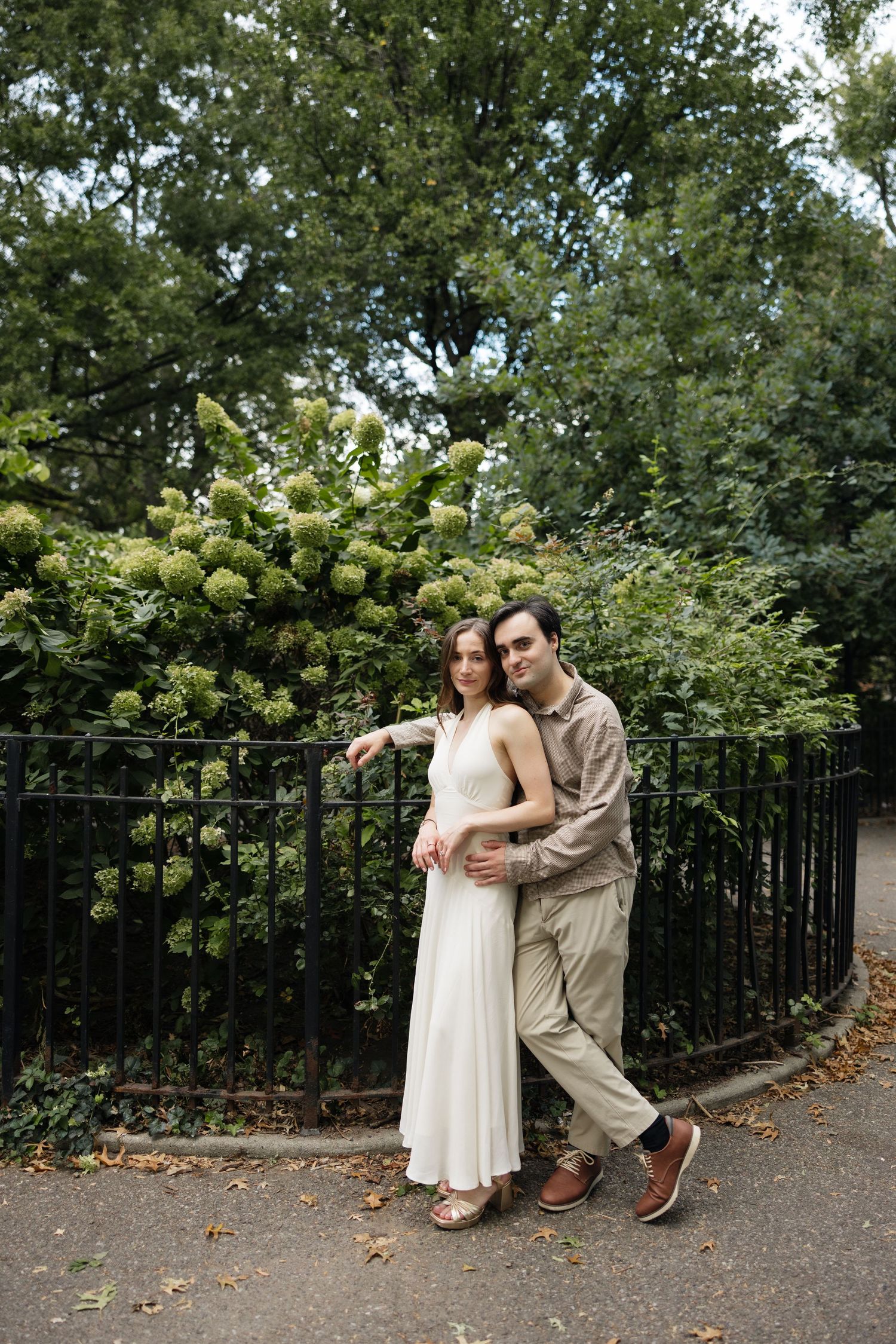 A couple poses together on a garden path with lush greenery and flowering bushes in the background.