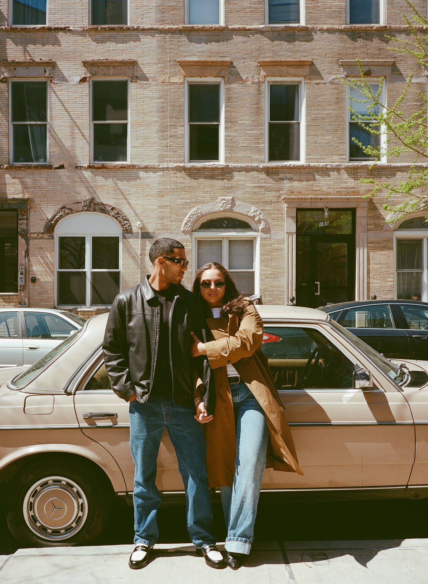 A vintage photograph shows a couple in casual wear standing next to a parked car on a city street with brownstone buildings.