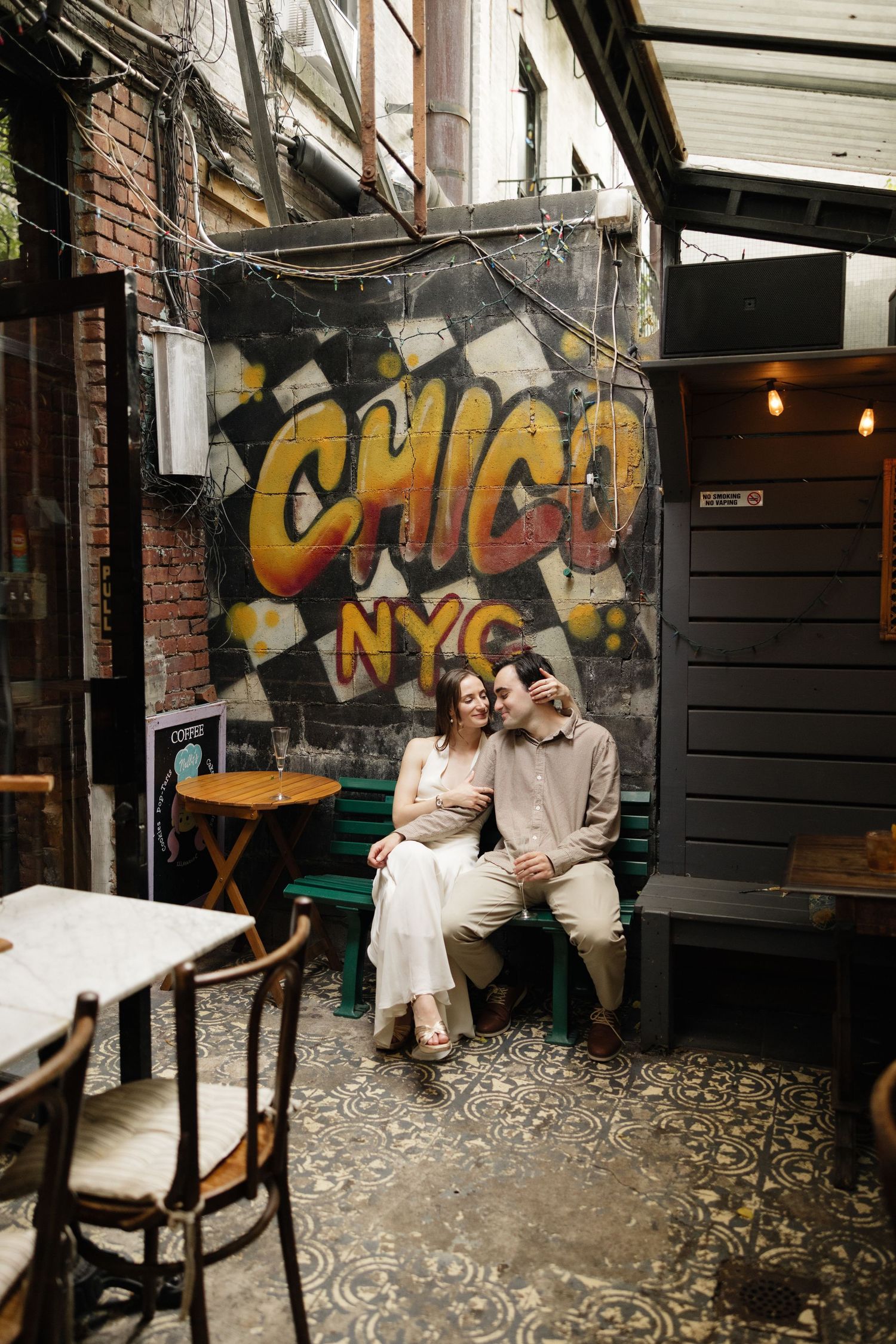 A couple sits together on a bench against a vibrant yellow graffiti wall reading 'CHICA NYC' in an urban cafe setting.