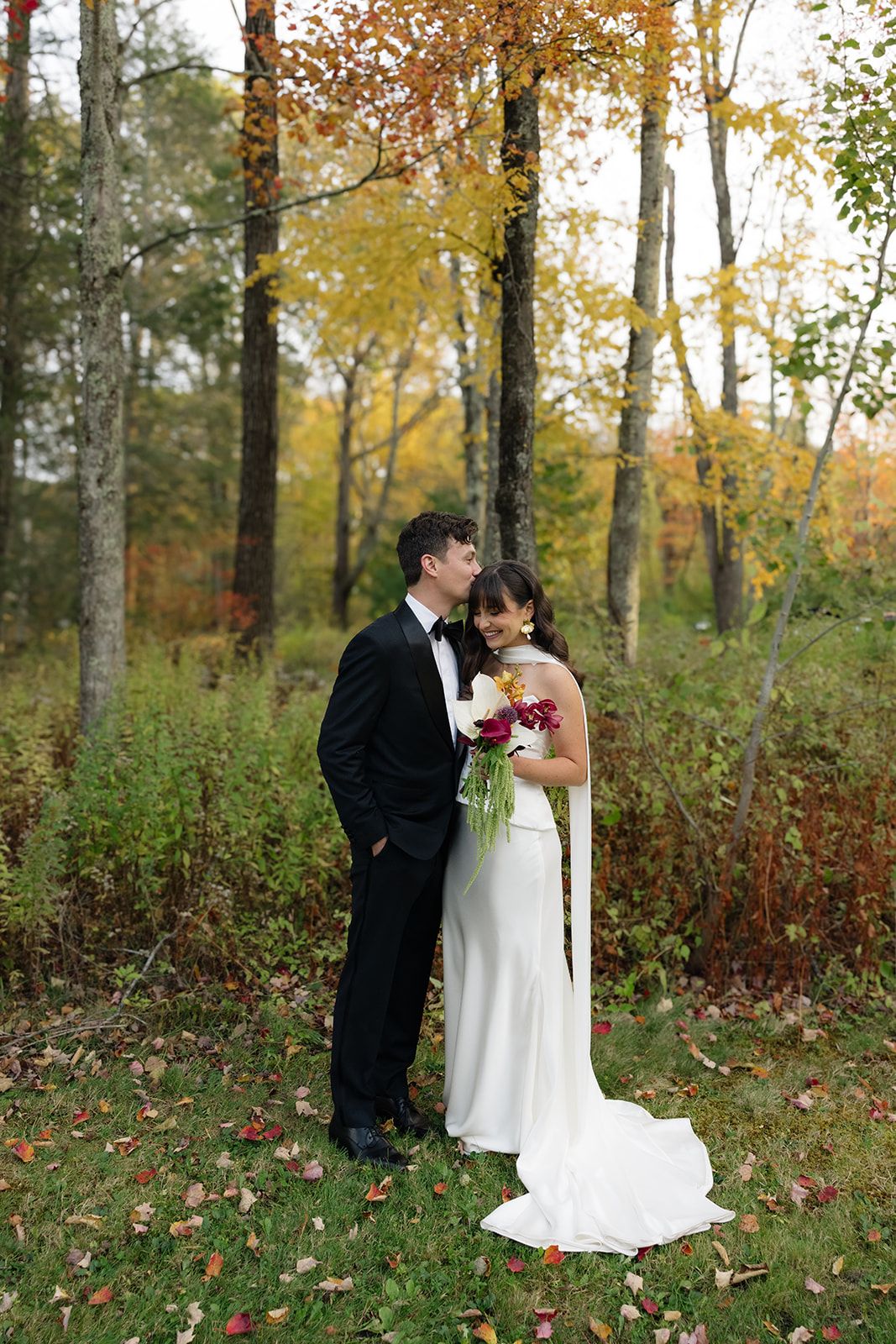 A couple shares a romantic moment in a wooded autumn setting with golden leaves and natural foliage.