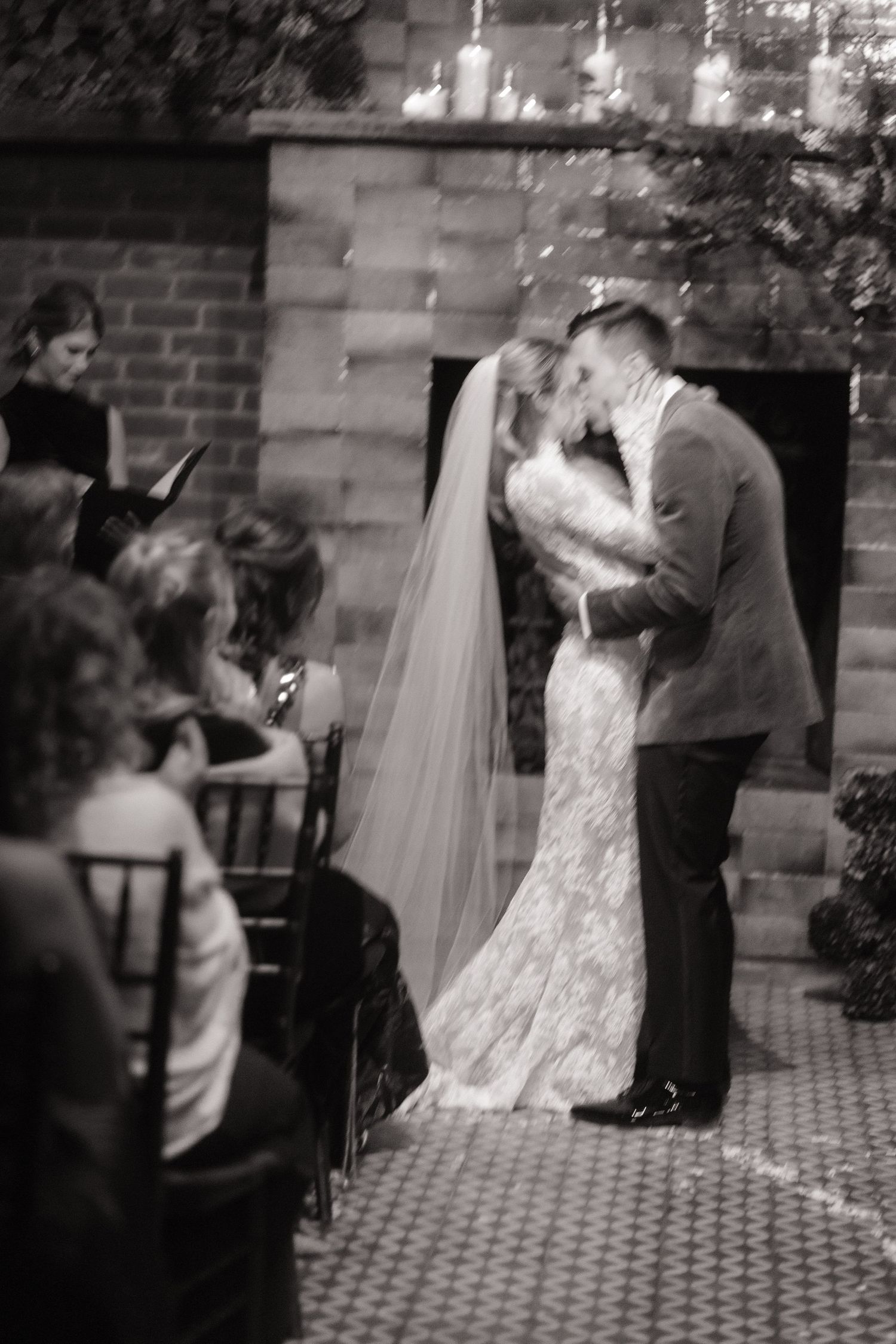 A romantic black and white wedding ceremony moment showing a couple's first kiss at the altar with guests looking on.