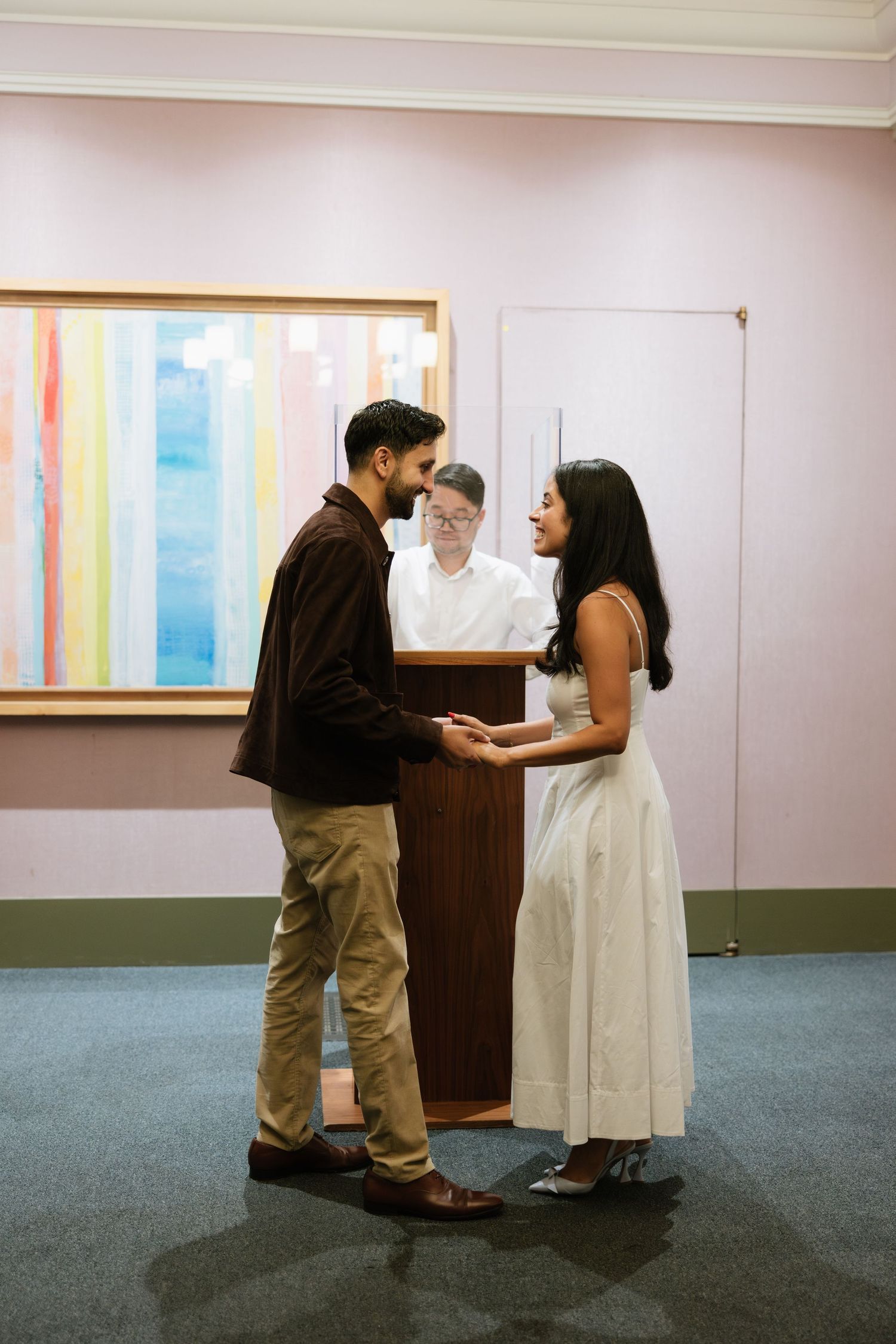 A couple stands at a registration desk in a gallery space with pastel rainbow artwork displayed on white walls.