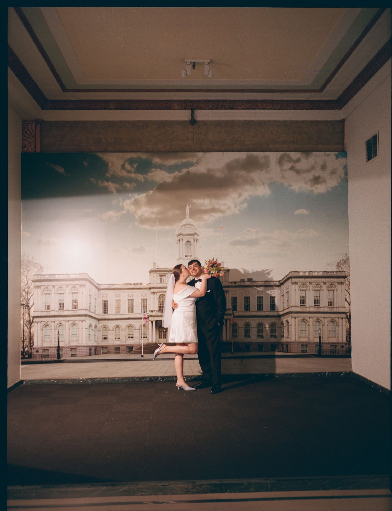 A couple embraces in front of a large backdrop featuring a grand historic building under dramatic clouds.
