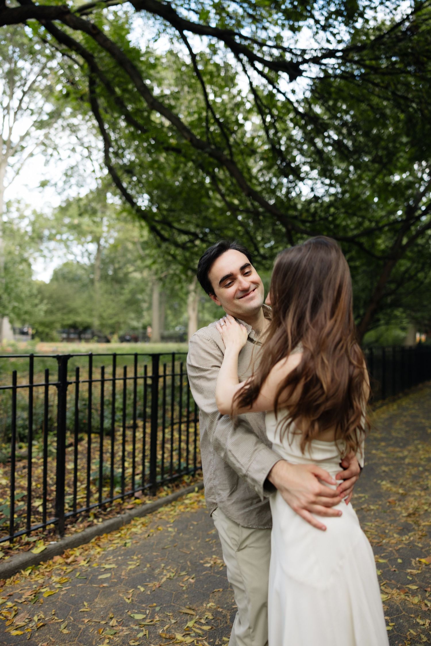 Romantic couple embraces and laughs together on a tree-lined path with autumn leaves and iron fence in the background.