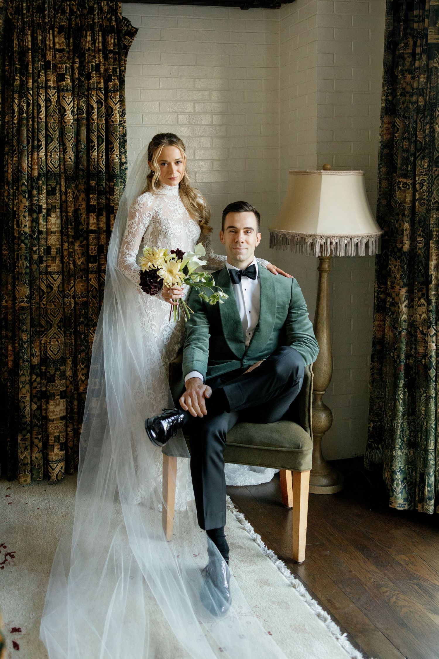 Elegant wedding couple poses in vintage-style room with green velvet chair and floral curtains.