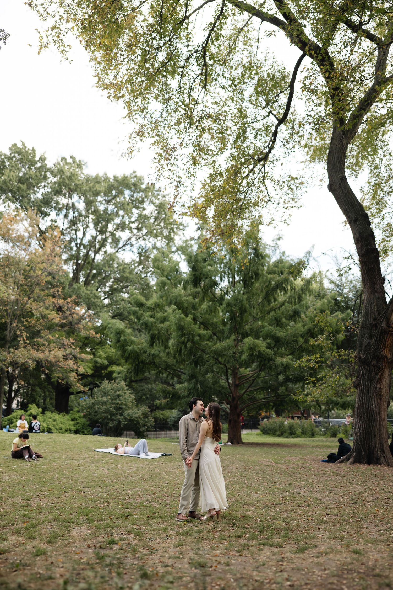 Couple walking together on grassy hill under autumn trees in Central Park with scattered people relaxing in background.