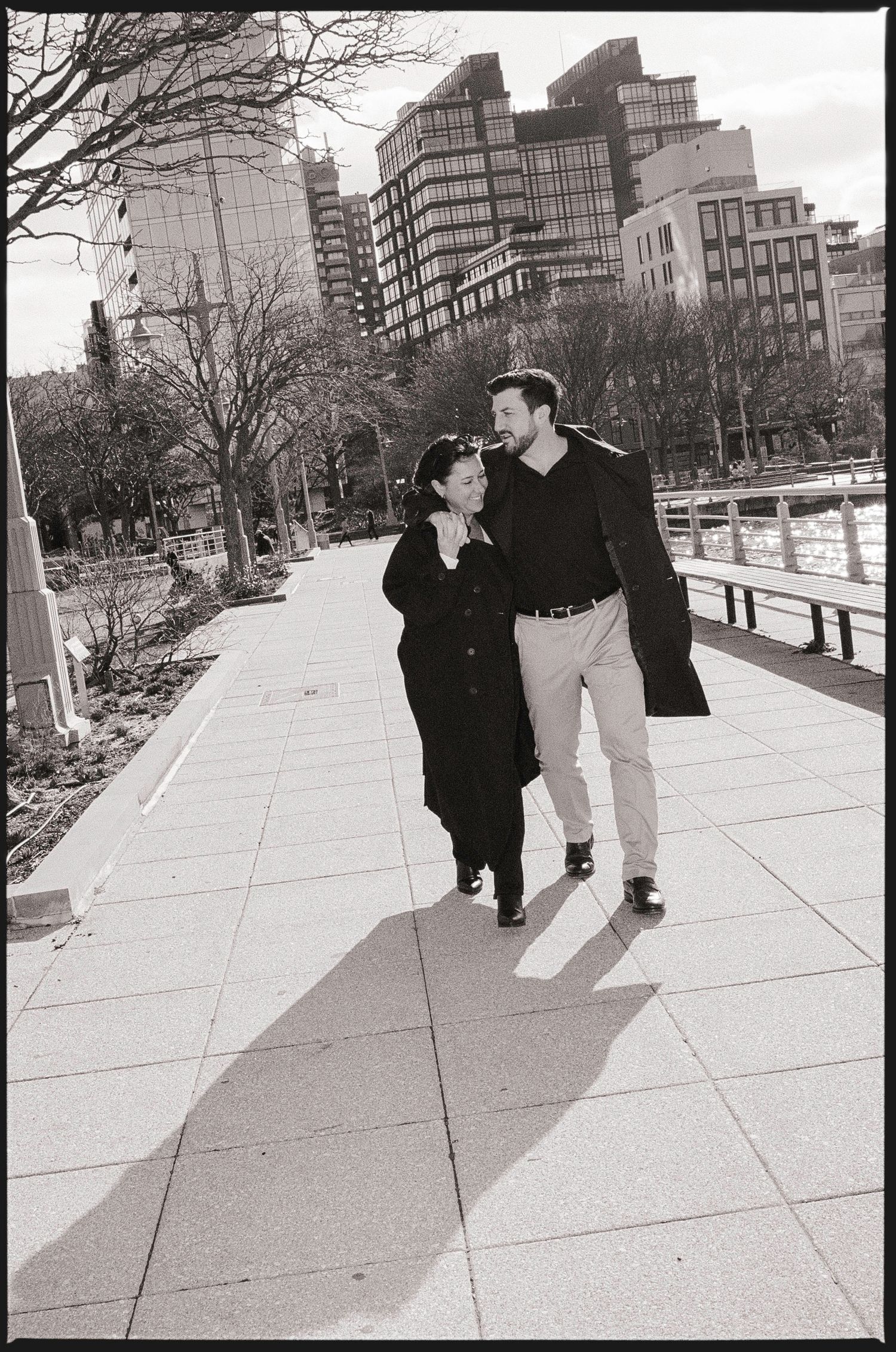 Black and white photo of a couple walking together on a city sidewalk, smiling and embracing near urban buildings.