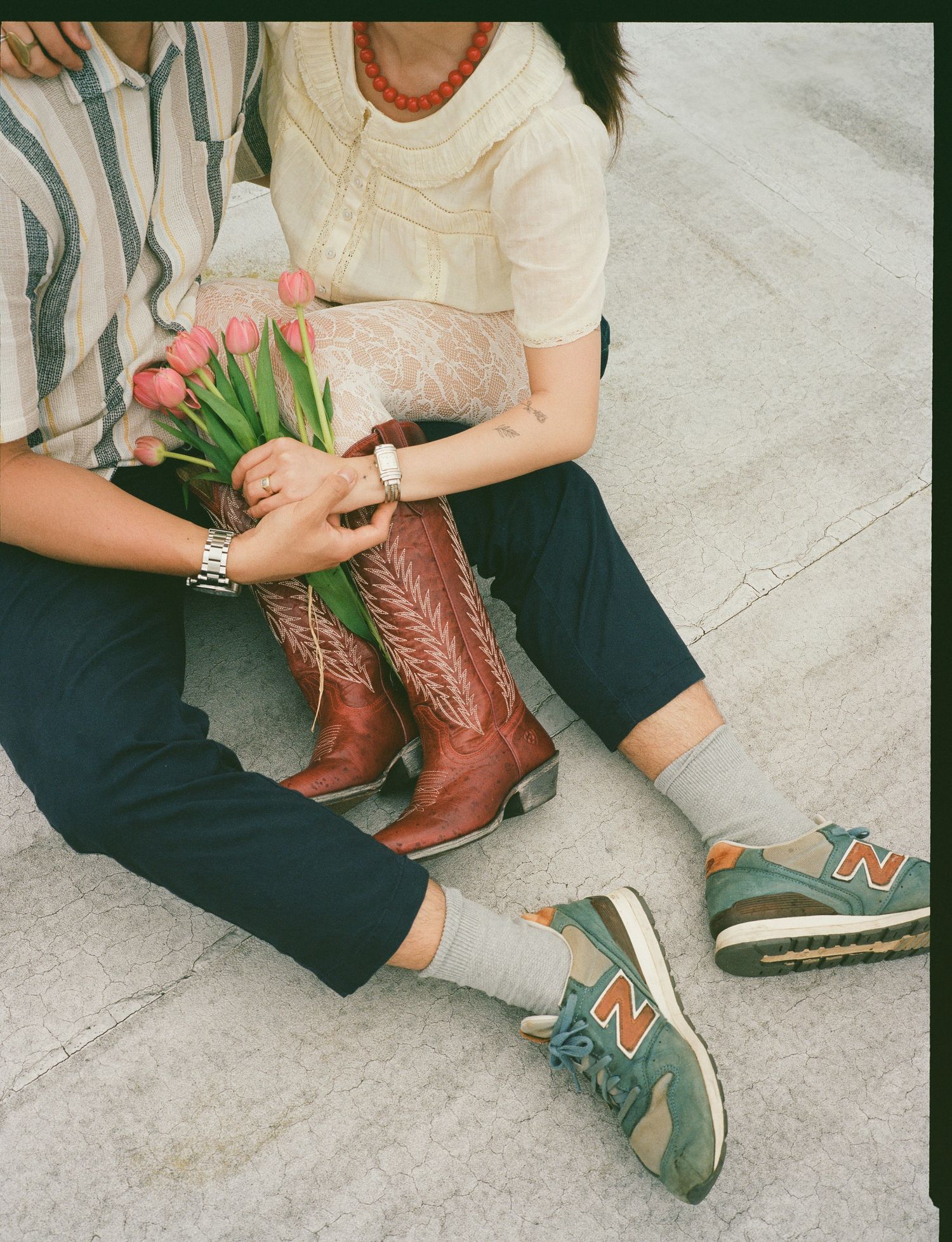 Person wearing brown cowboy boots and sneakers sits on concrete holding pink tulips in a casual spring fashion scene.