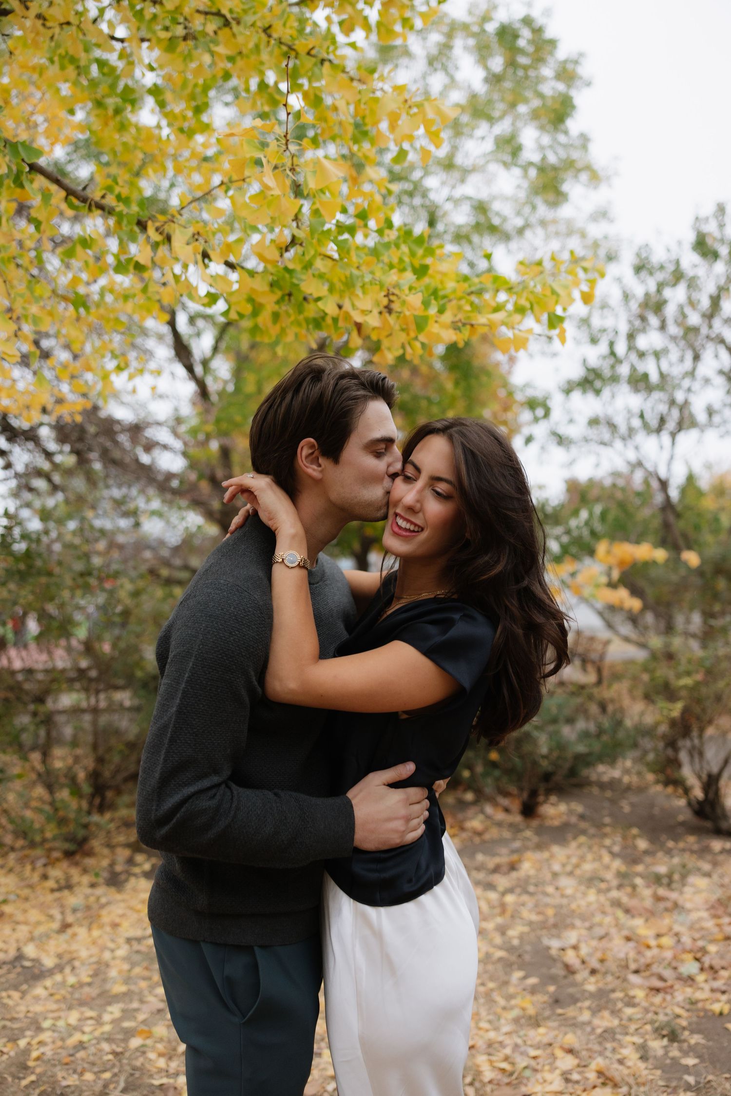 A couple embraces and laughs together during an autumn photoshoot with yellow leaves in the background.