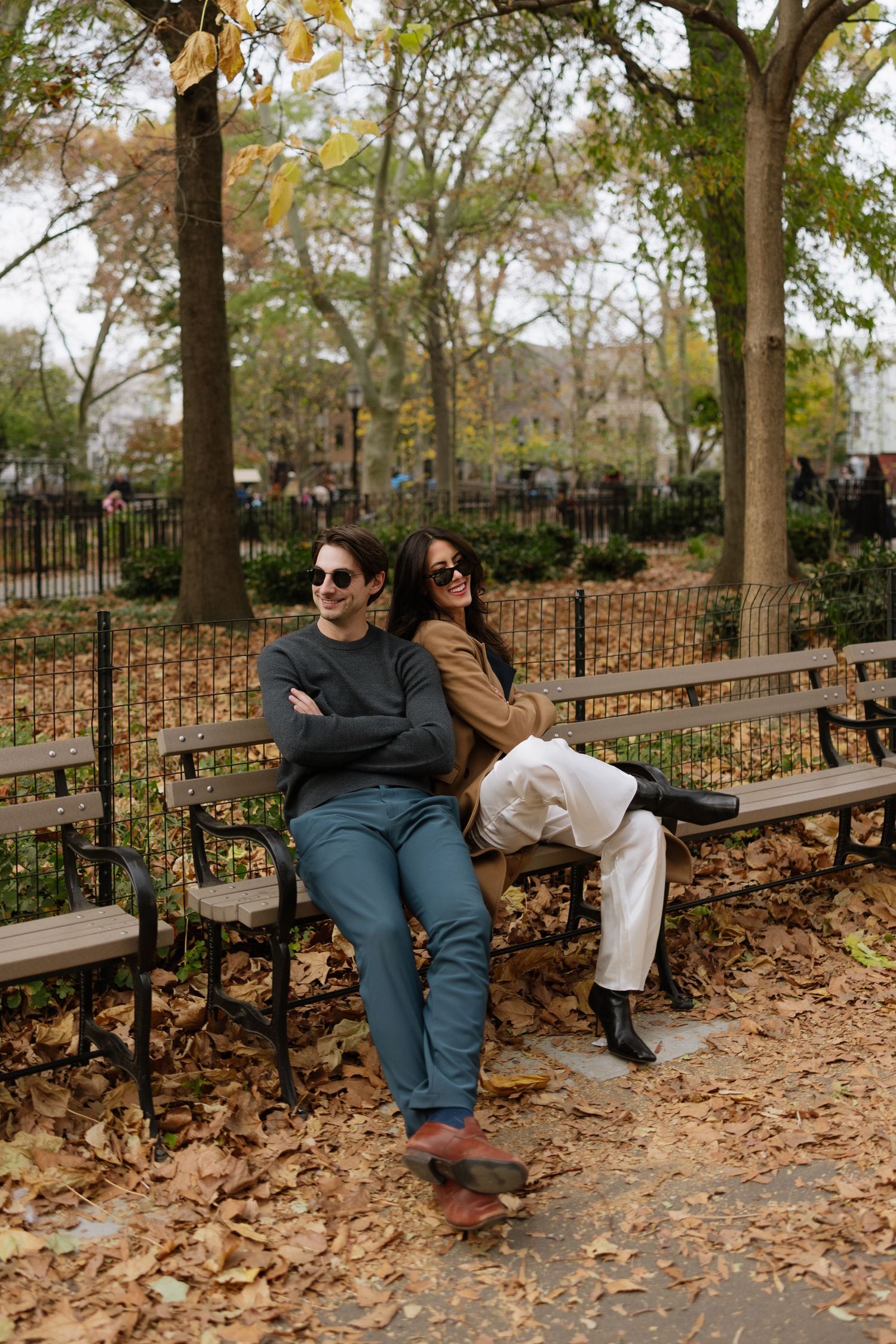Two people sit together on a park bench surrounded by autumn leaves and trees in an outdoor setting.