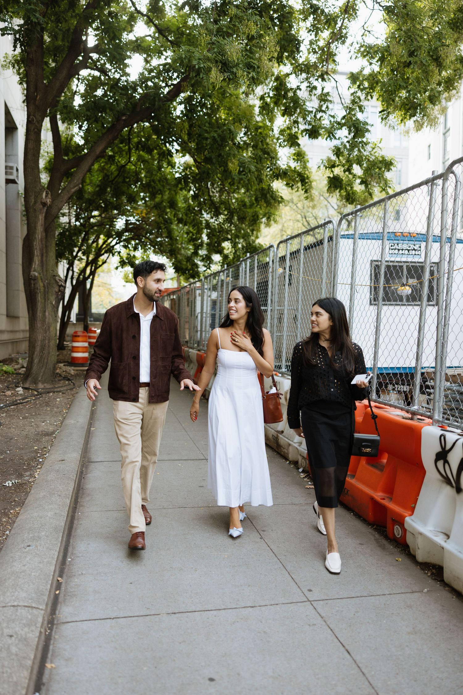 Three people walking on a sidewalk in casual attire with trees and a chain-link fence in the background.