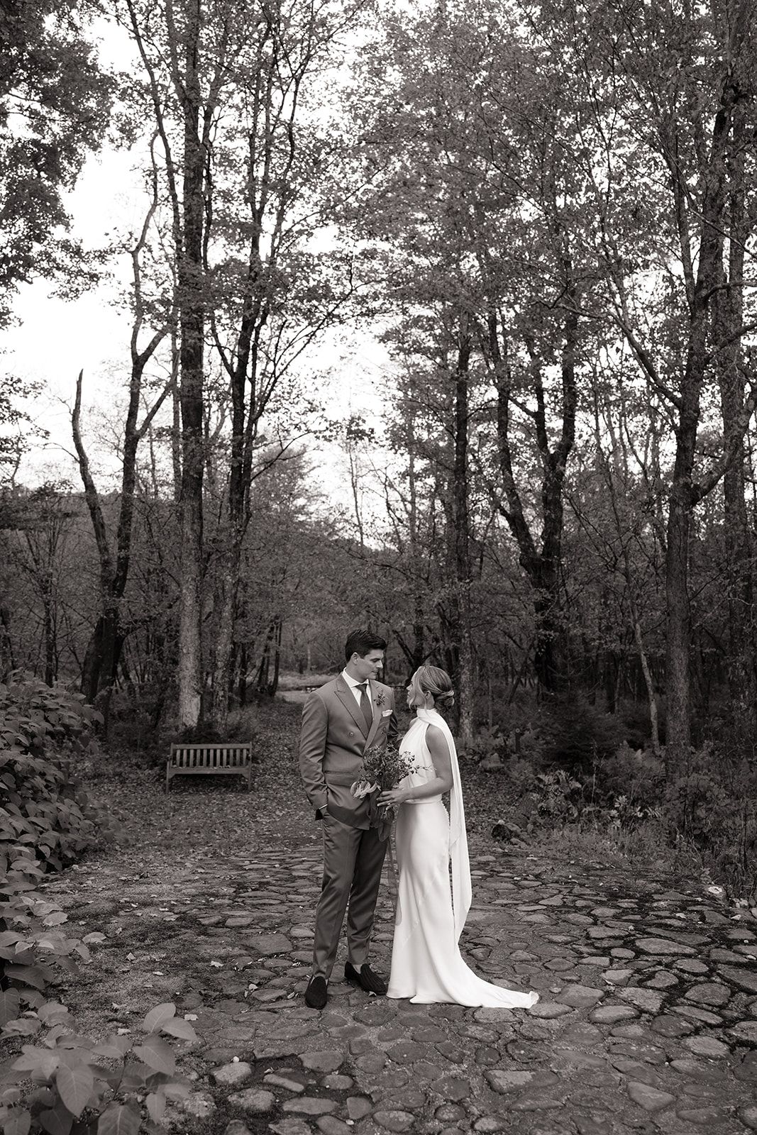 A romantic black and white photo series showing a couple in wedding attire walking along a stone path through the woods.