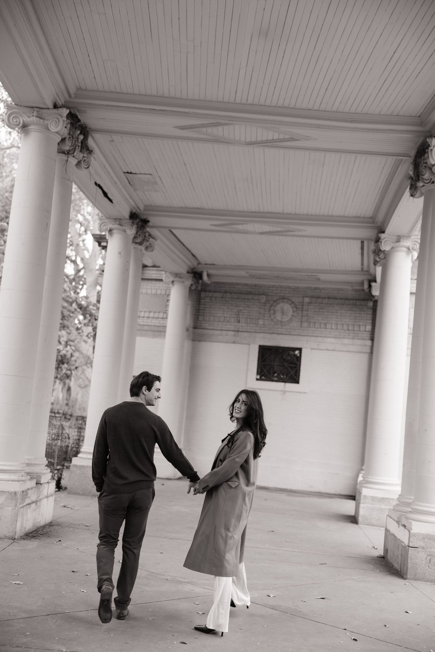 Two people walk through a minimalist architectural corridor with white columns and ceiling creating a dramatic perspective.