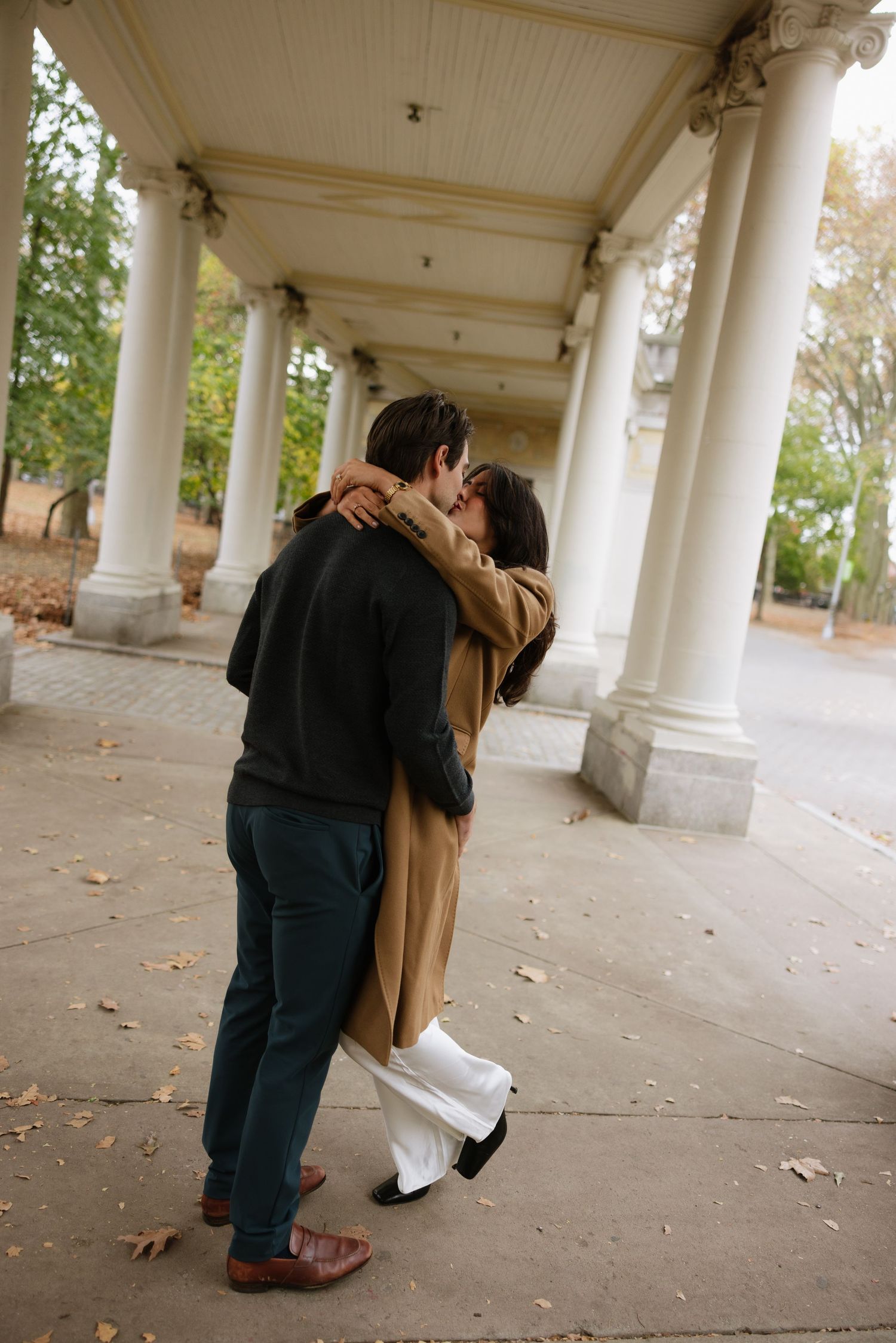 A romantic couple embraces in a colonnade walkway with white pillars and fallen autumn leaves on the ground.