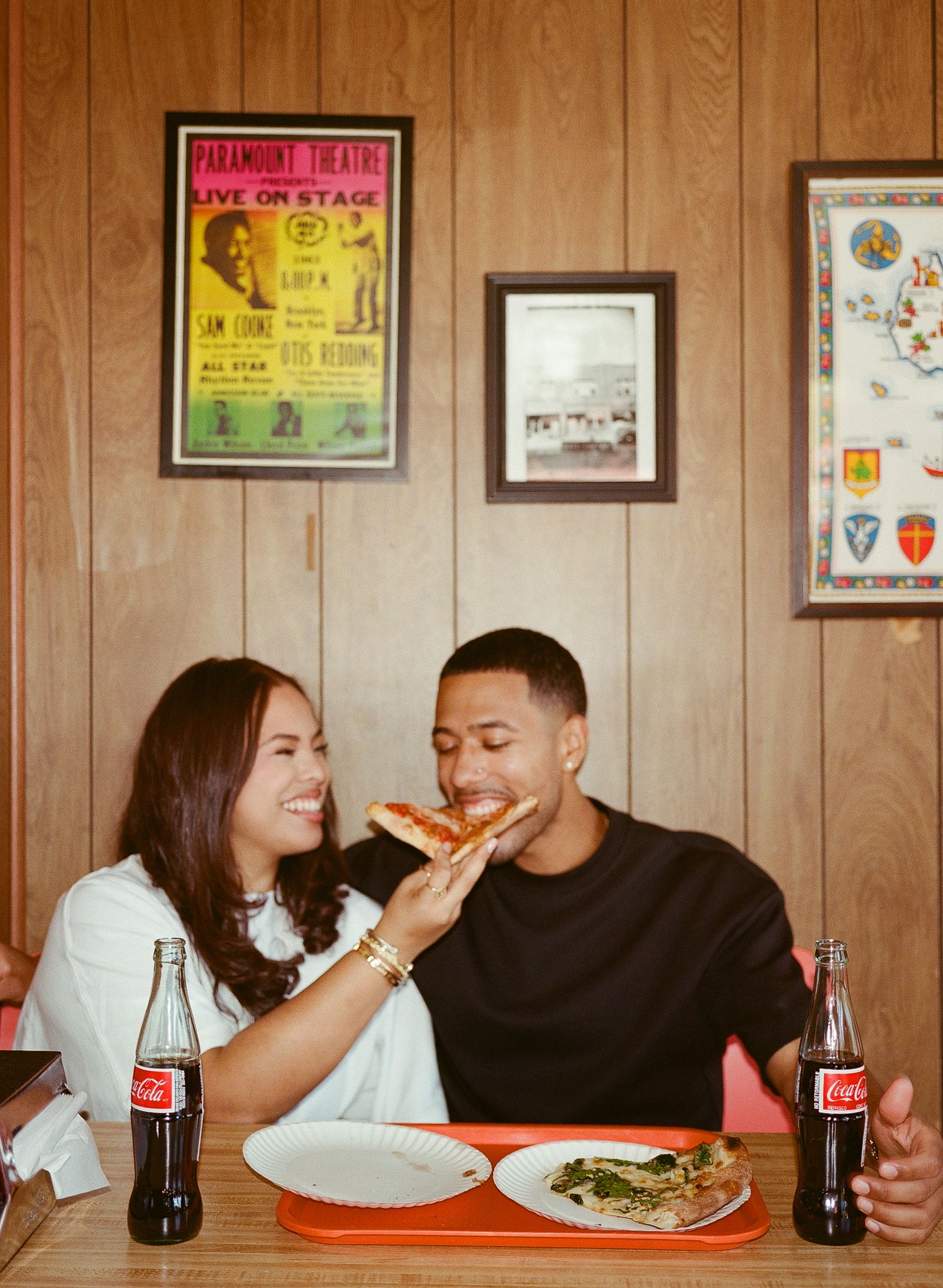 A couple shares pizza and drinks while dining together at a casual restaurant with vintage posters on wood-paneled walls.