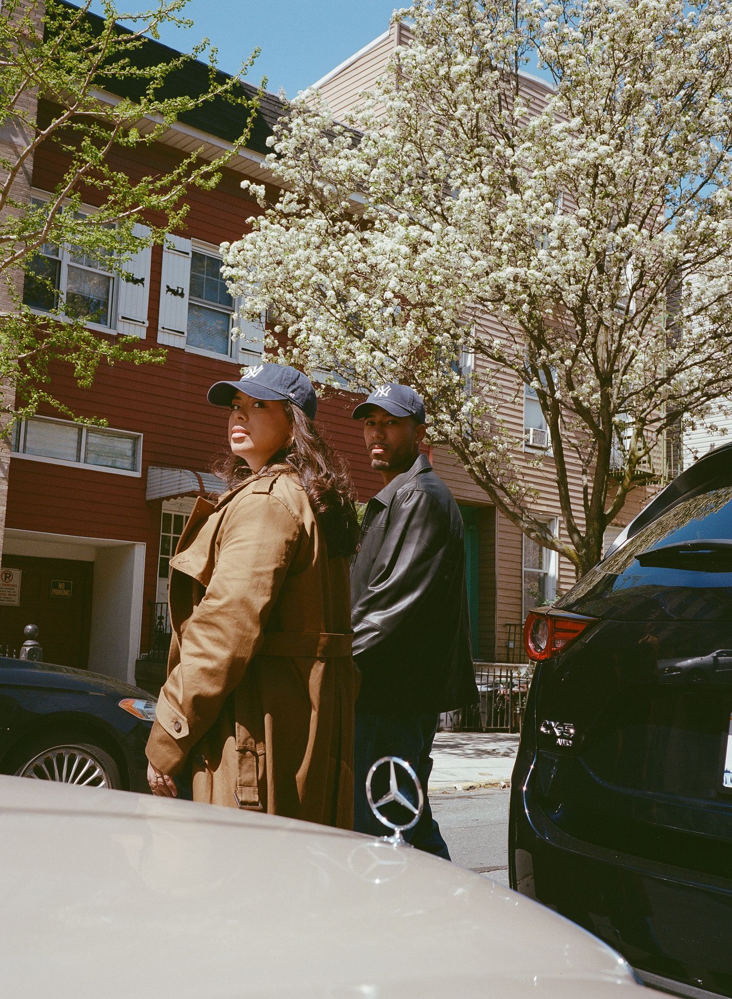 Two people stand next to parked cars on a spring day with blooming cherry trees and apartment buildings in the background.