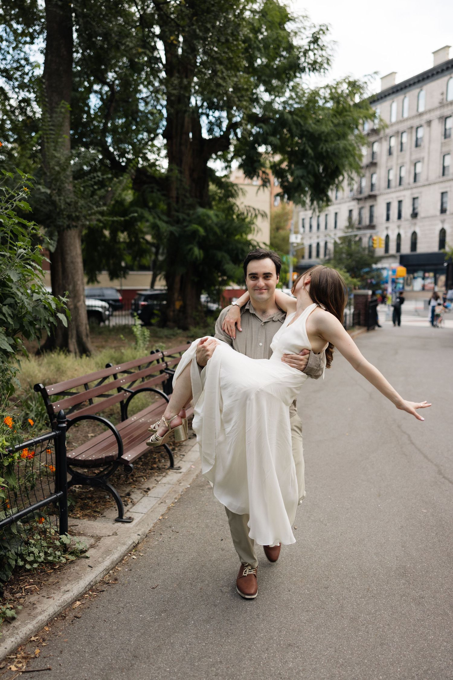 A person in a white dress spins playfully on a park path with benches and buildings in the background.