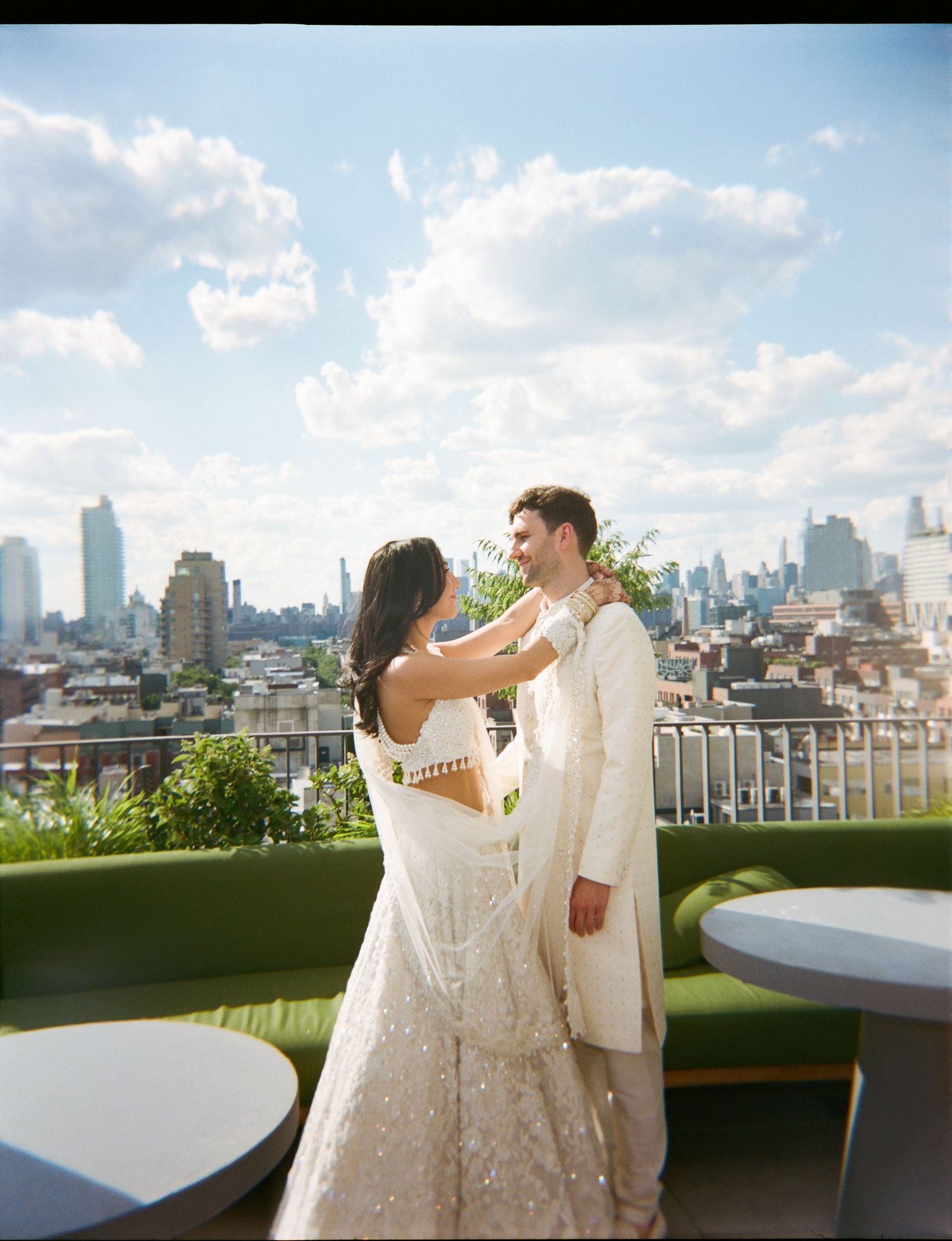Couple in white wedding attire sharing a romantic moment on a rooftop with a city skyline backdrop.