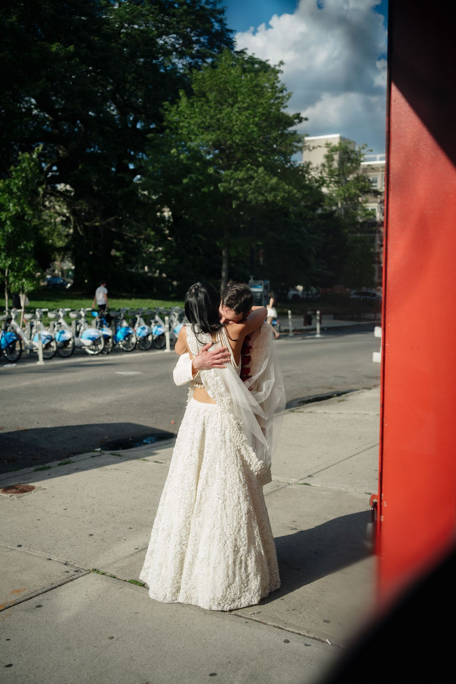 Two brides in white wedding attire embrace on a sunny urban sidewalk near a bike share station.