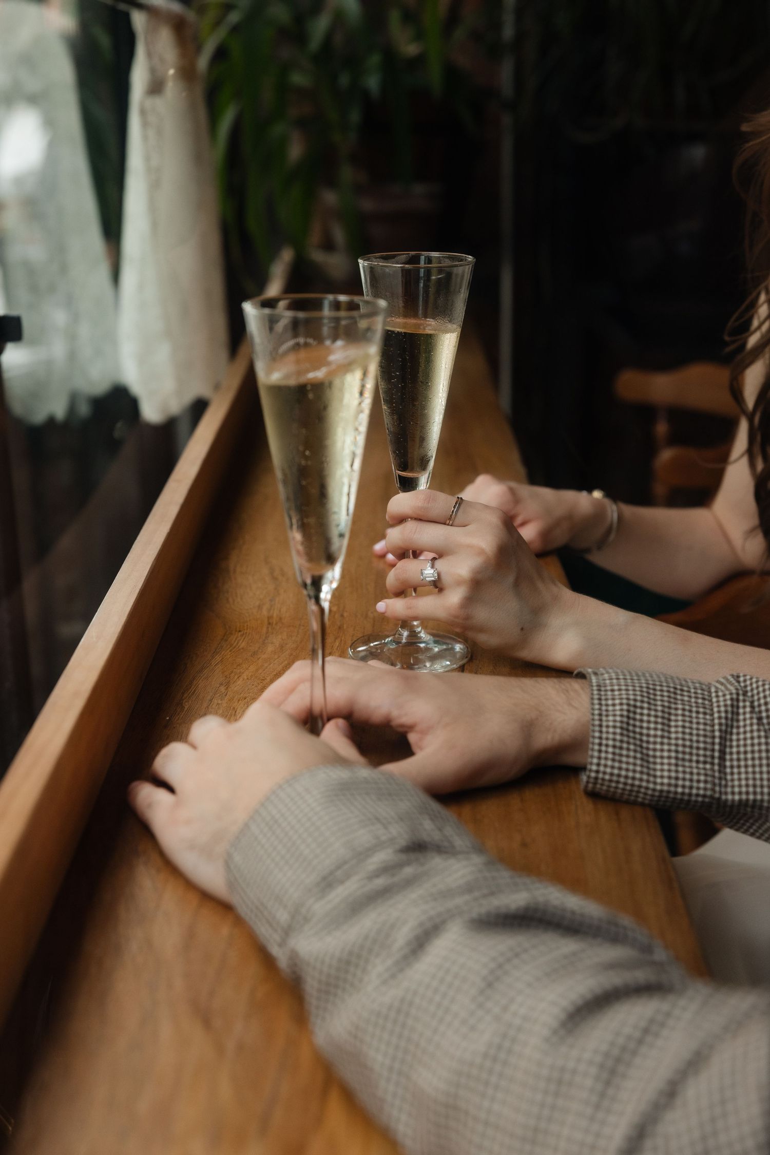 Two glasses of champagne on a wooden bar counter with hands reaching for them in an intimate setting.