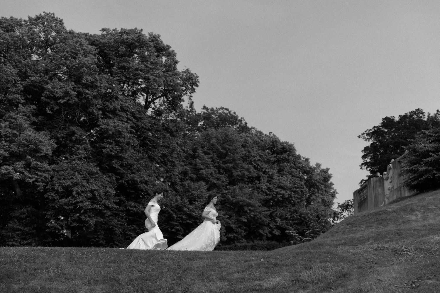 Two brides in white gowns walk across a grassy hill surrounded by lush trees in a black and white wedding photo.