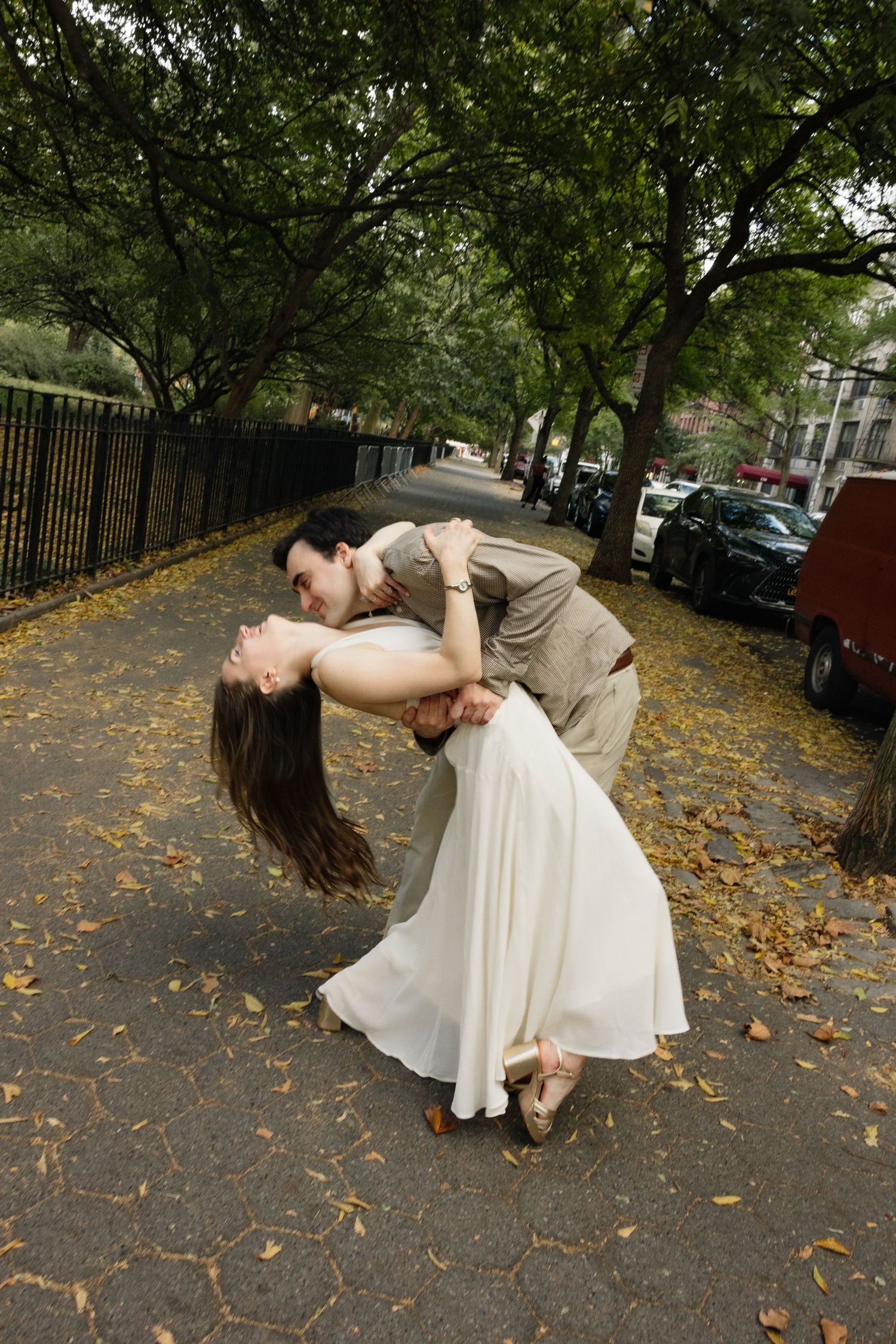 A couple in formal attire performs a romantic dance dip pose on a tree-lined path covered in autumn leaves.