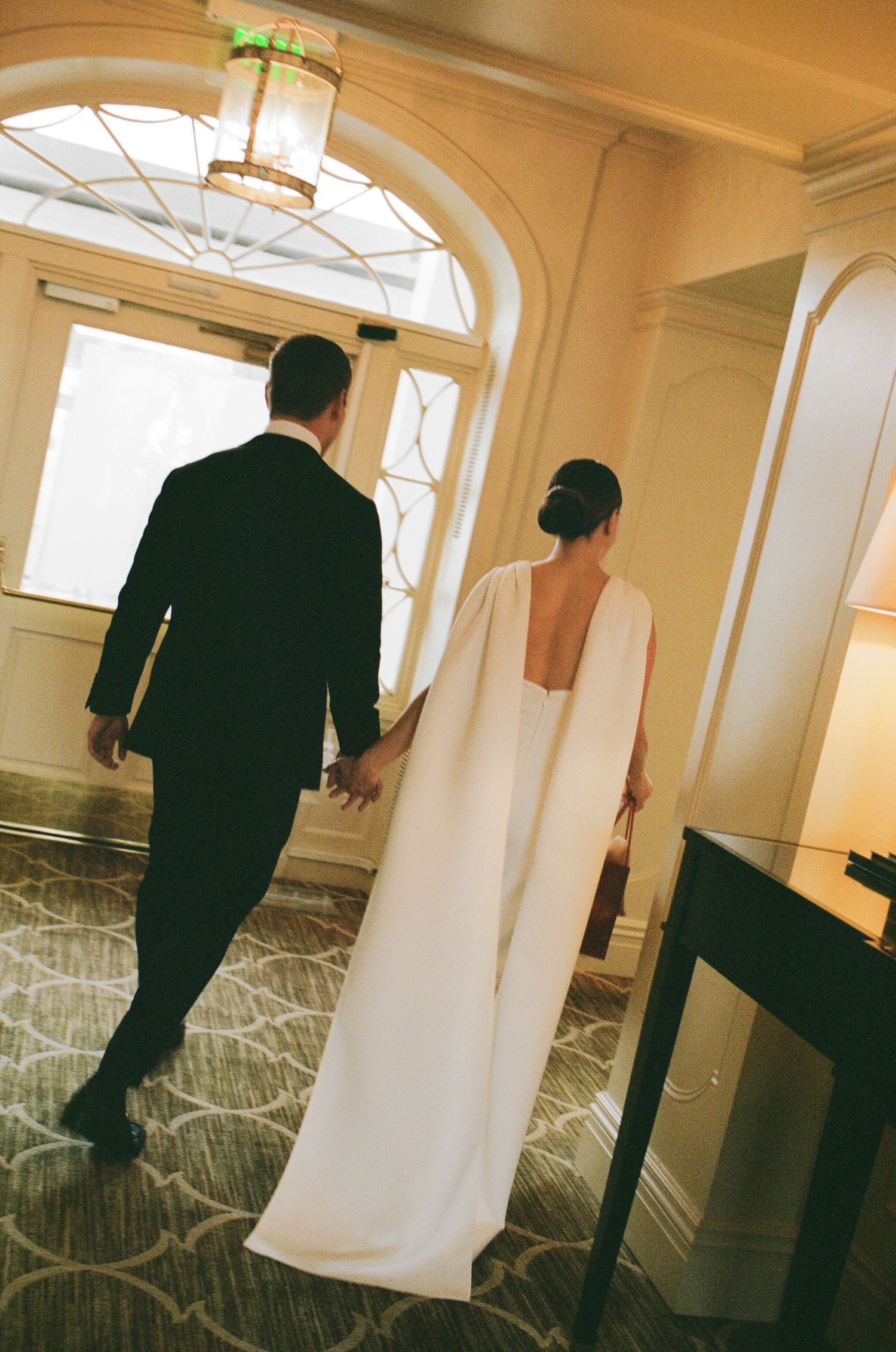 A bride in a white cape gown and groom in black suit walk hand in hand through an elegant hotel corridor.