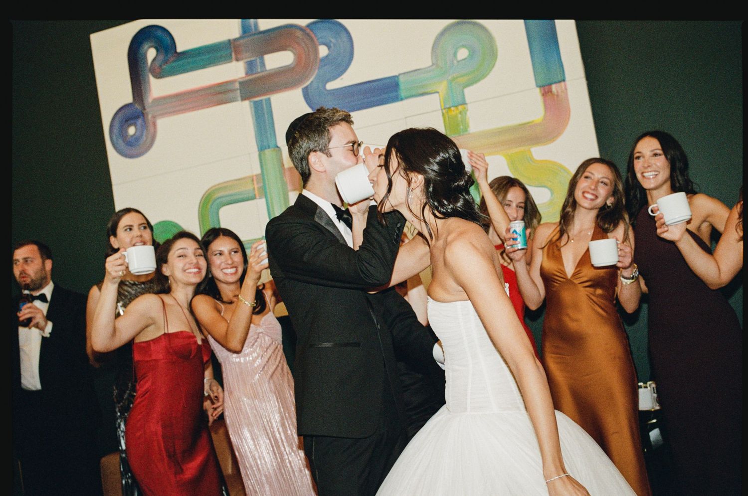 Bride and groom share a kiss surrounded by bridesmaids holding white mugs at a colorful wedding reception.