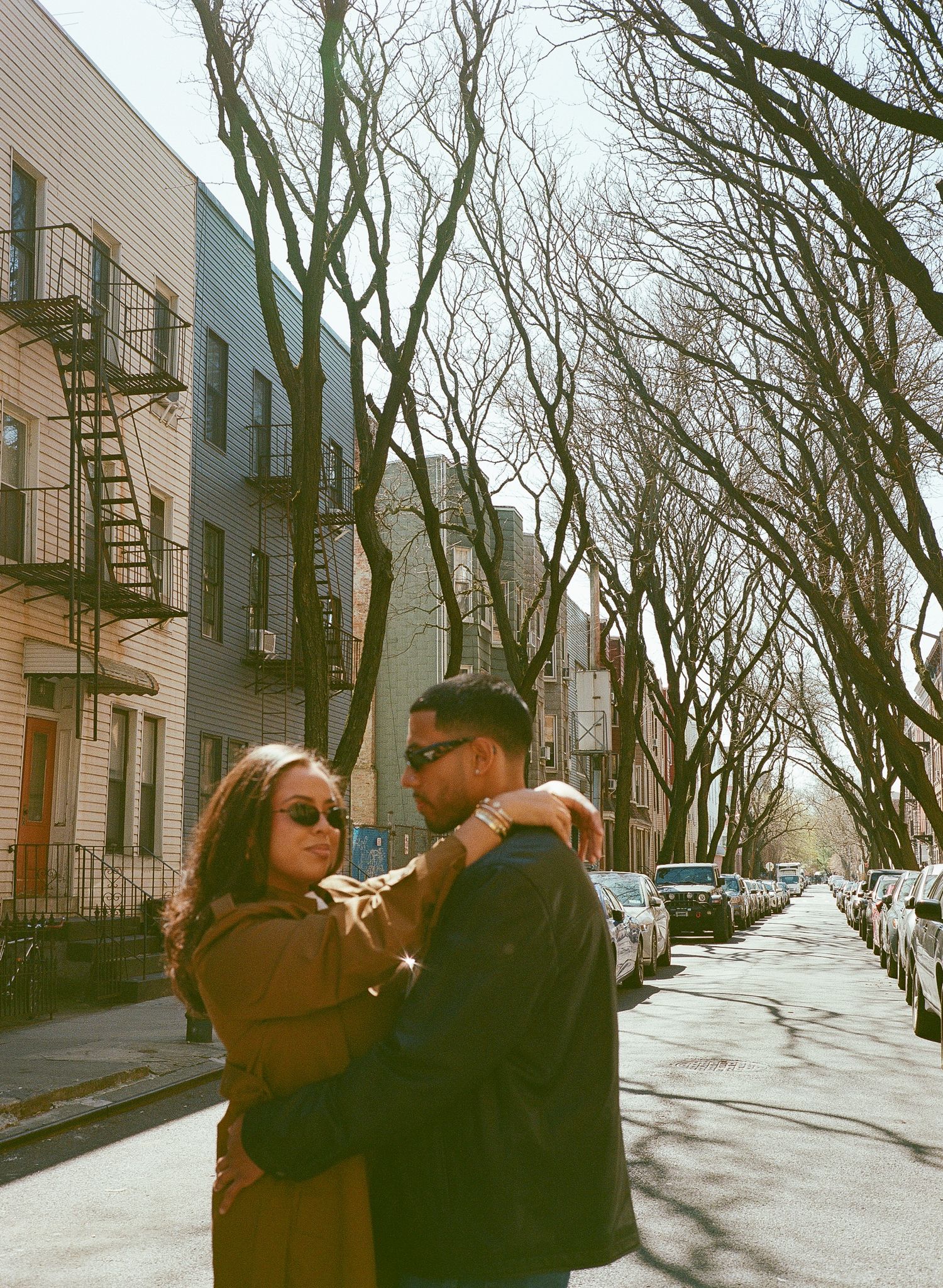 A couple embraces on a tree-lined street with brownstone buildings and fire escapes on a sunny winter day.