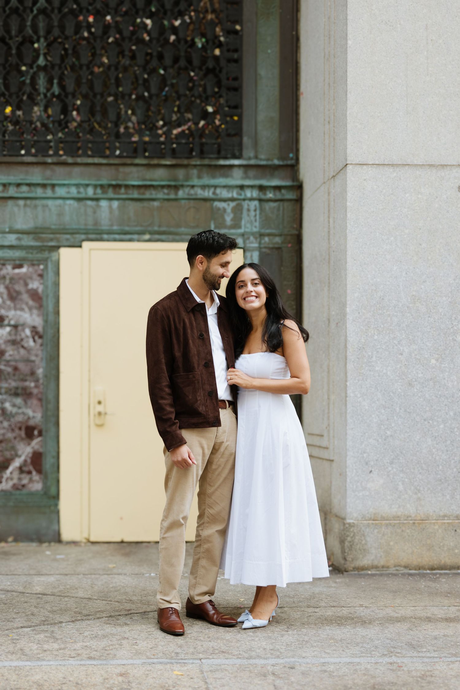 A couple in contrasting brown and white outfits share an intimate moment against a stone wall exterior.