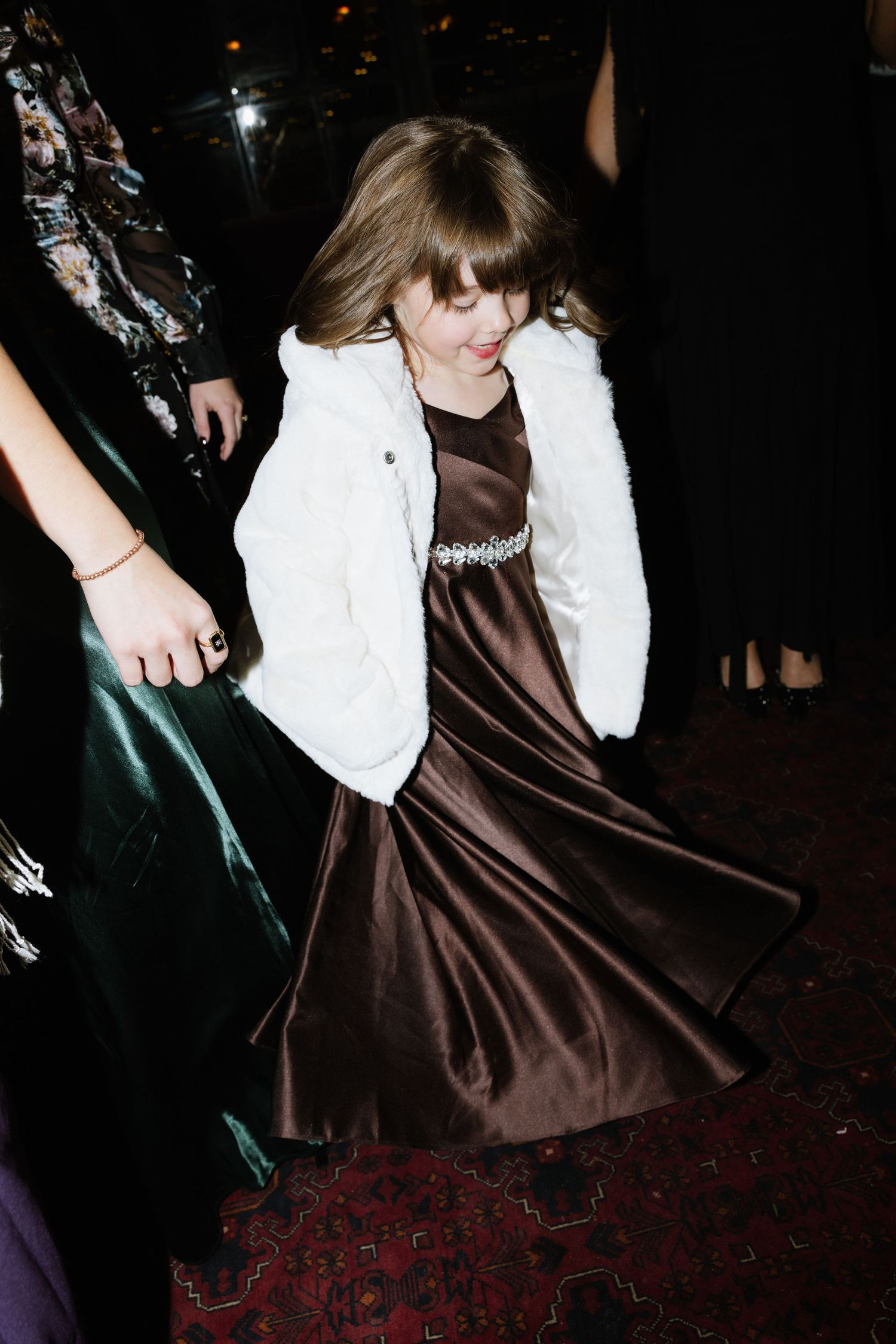 A person in a white top and brown satin dress spins and twirls on a patterned red carpet in a dark room.