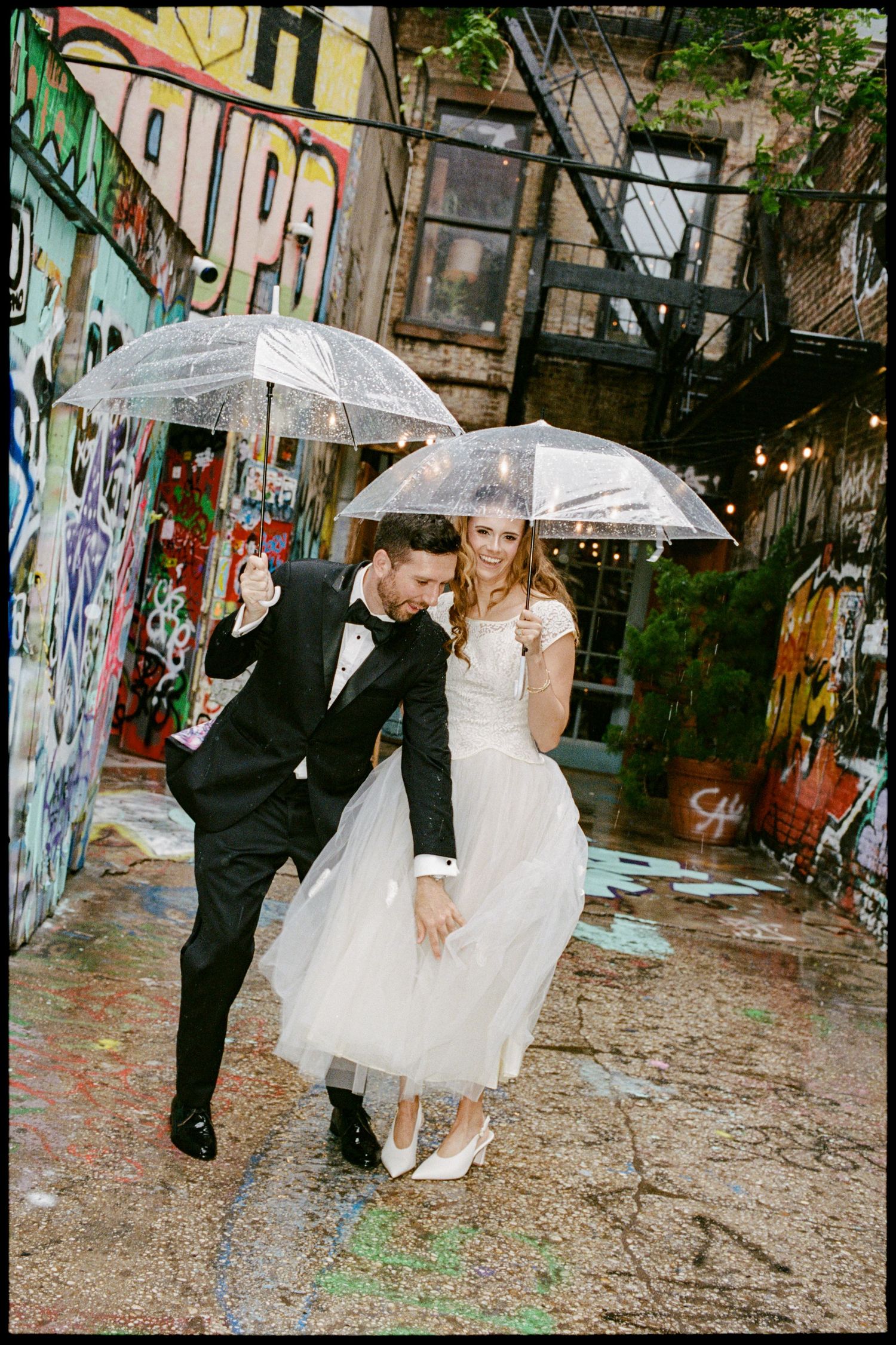 Bride and groom dancing in the rain with clear umbrellas in a colorful graffiti alley on their wedding day.