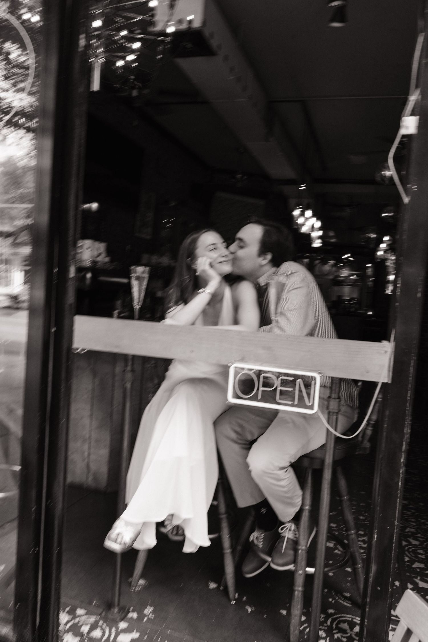 A couple shares a romantic kiss in front of a storefront with an 'OPEN' sign visible in the window.