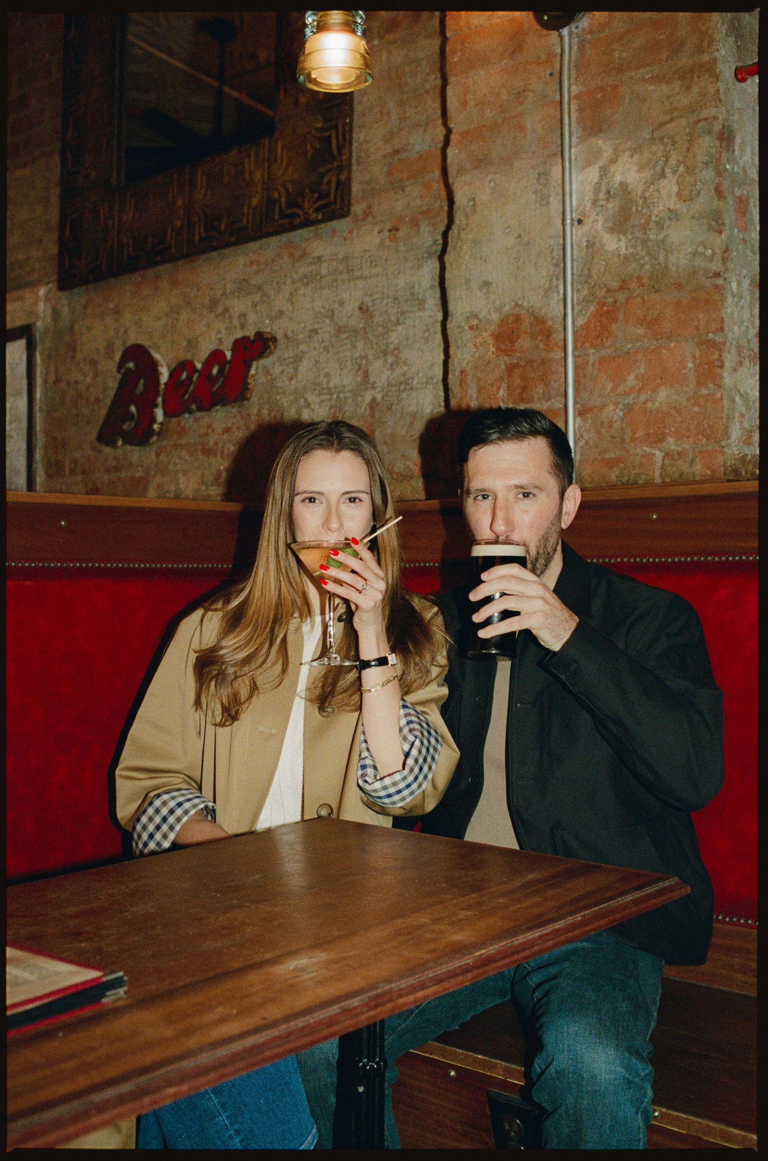 Couple sitting at a wooden bar table in a cozy pub, woman holding cocktail, man drinking dark beer.