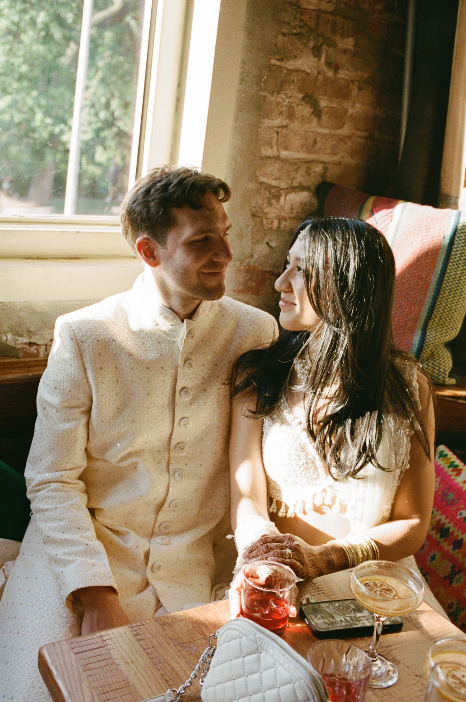 A couple in elegant white attire sharing a romantic moment at a sunlit table with drinks.