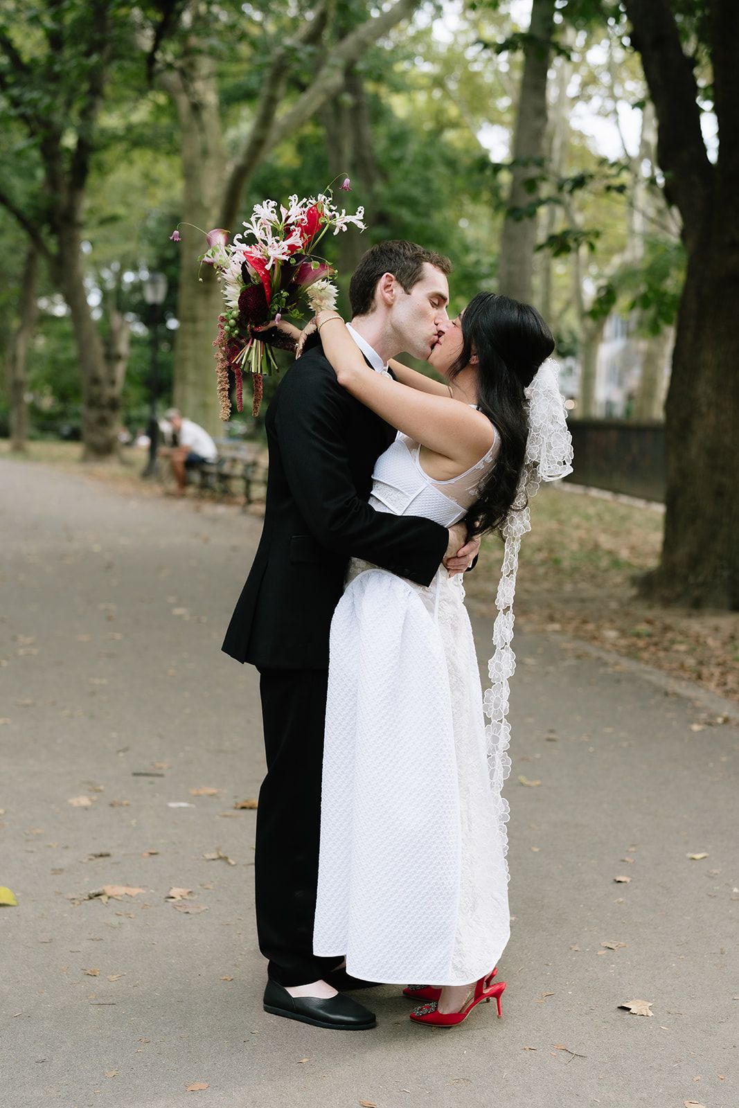 A couple in formal wedding attire shares a romantic kiss in a tree-lined park setting with fallen autumn leaves.