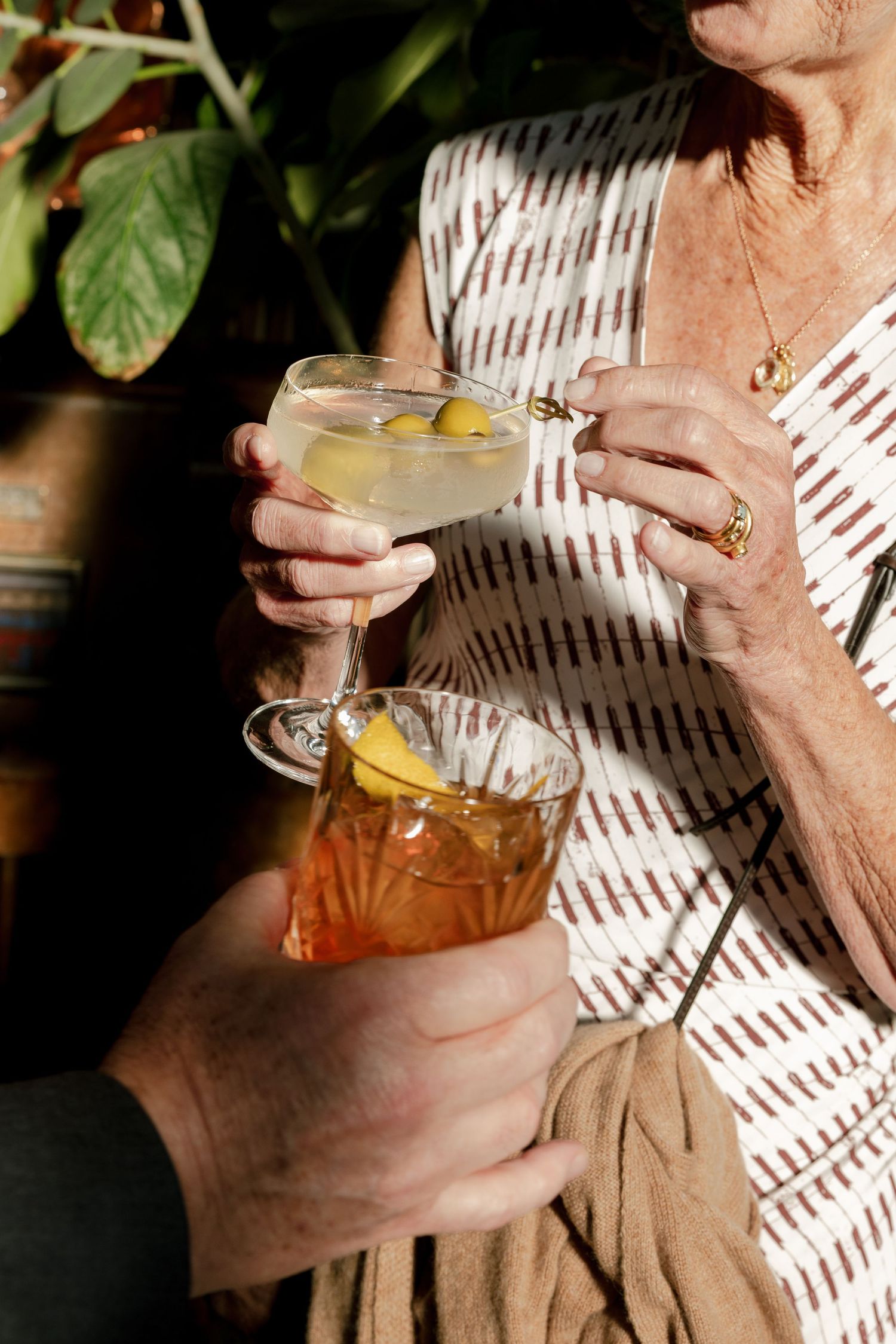 Elderly woman holding a martini with olives clinking glasses with an old fashioned cocktail outdoors.