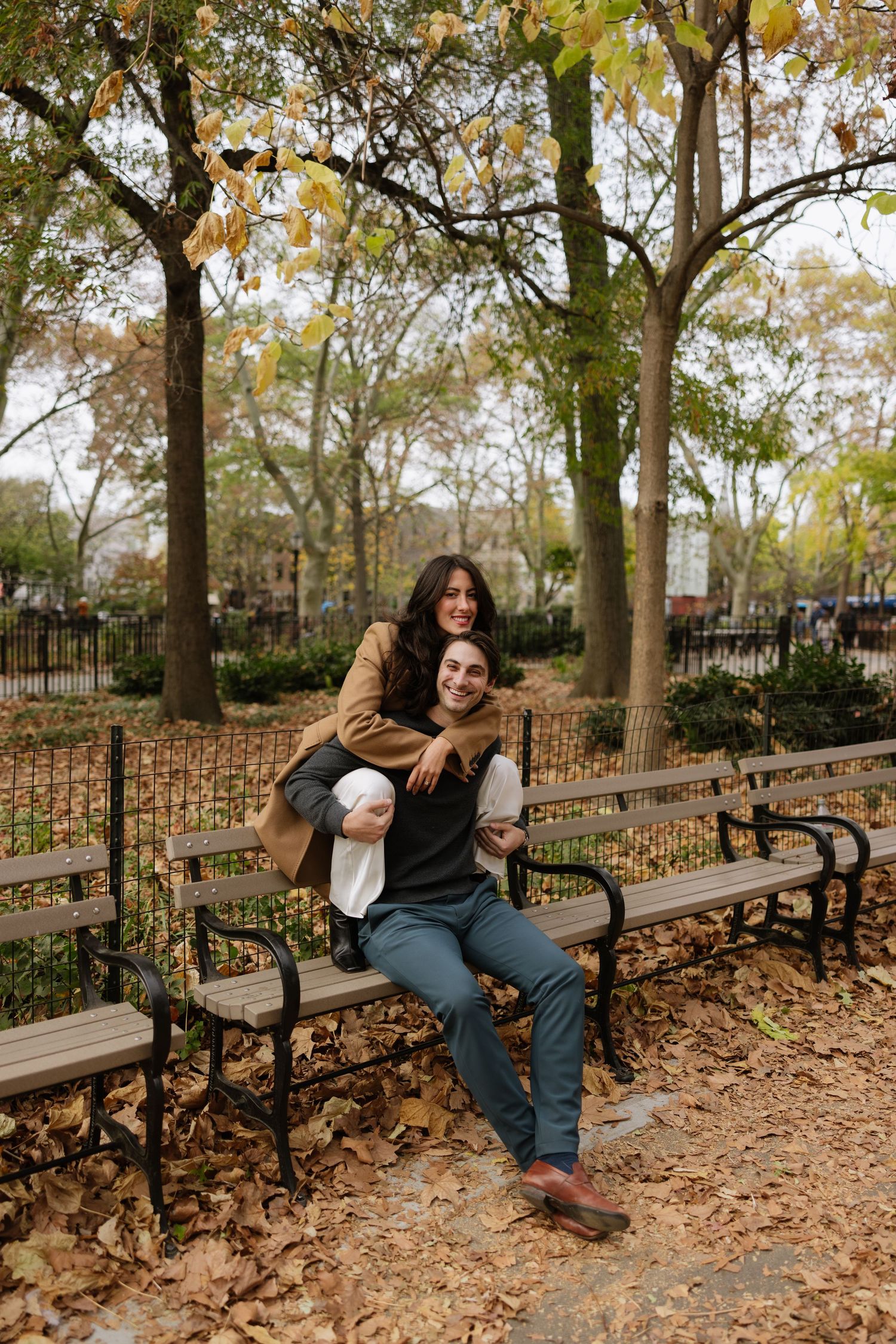 Two people sitting together on a wooden park bench surrounded by autumn leaves and trees in a tranquil setting.