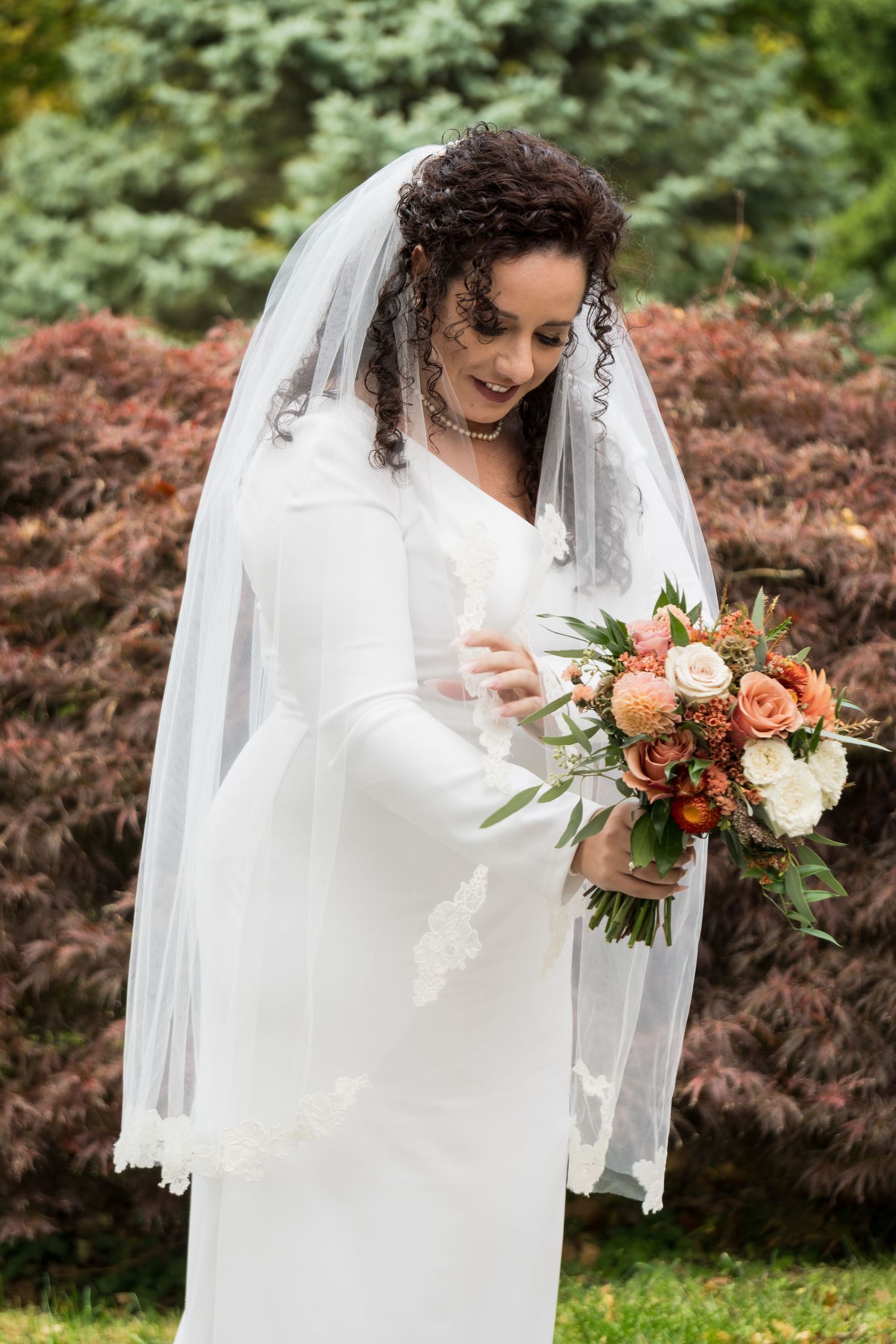 A bride in a flowing white dress holds a peach and white floral bouquet against a backdrop of autumn foliage.