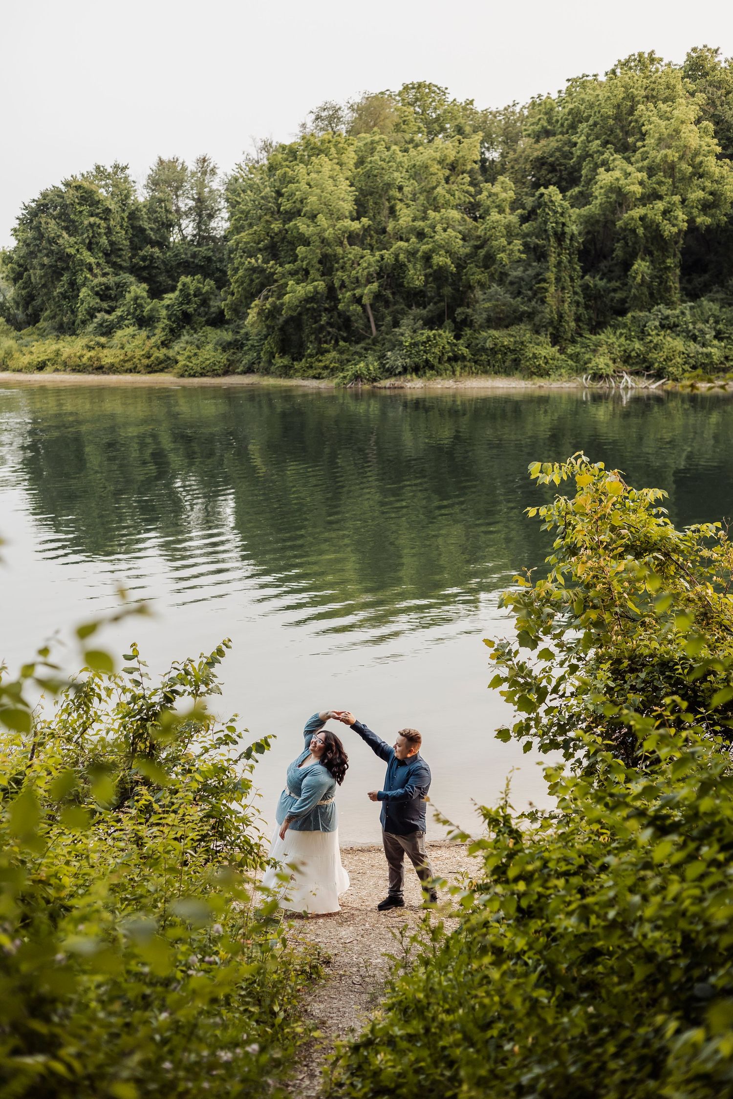 Two people share a romantic moment by a serene lake surrounded by lush greenery.