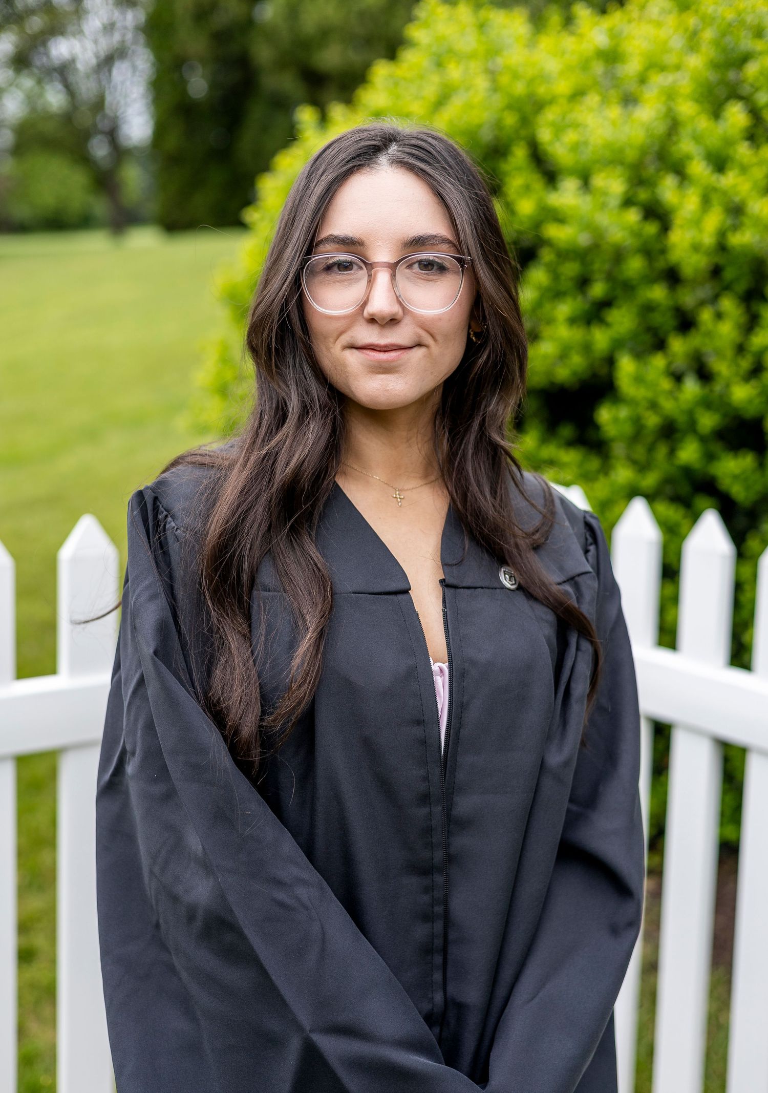 Portrait series of person in black graduation gown against white picket fence and green bushes.