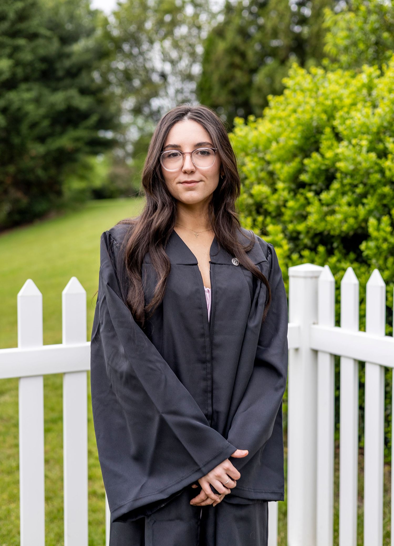 Graduate in black cap and gown poses by white picket fence with green landscaping background.