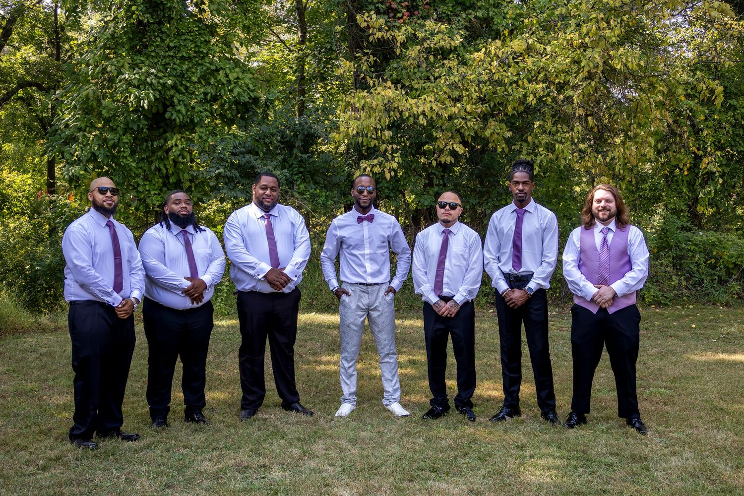 Wedding party members in purple dresses and white shirts lined up outdoors against green foliage.