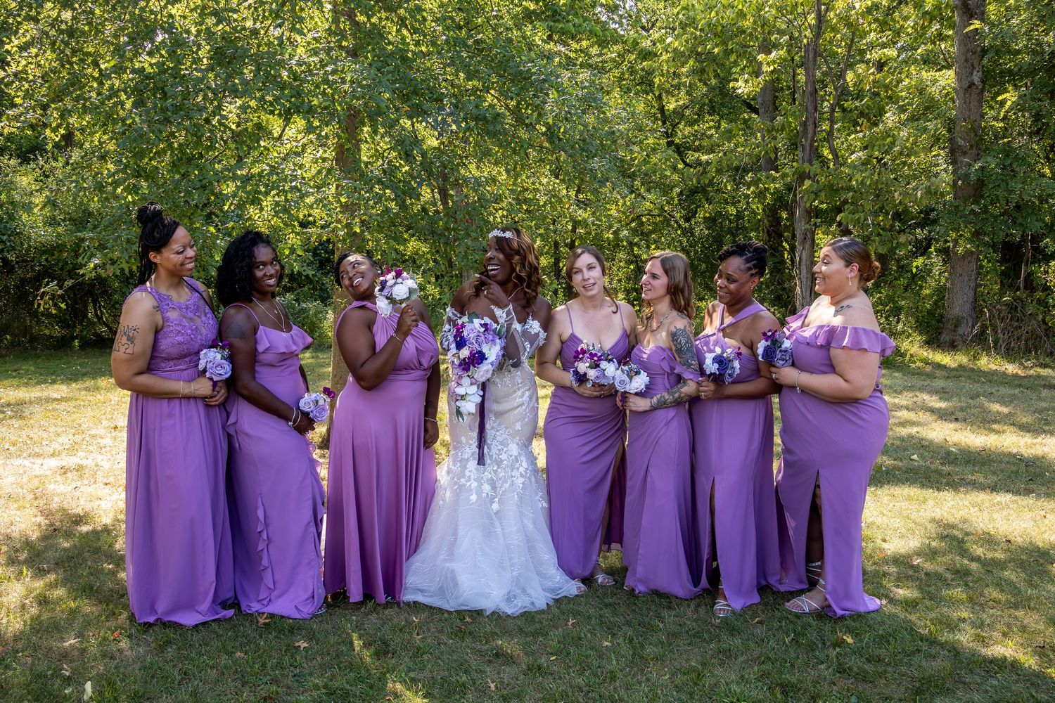 Wedding party wearing purple and white attire posed together on grass with trees in background.