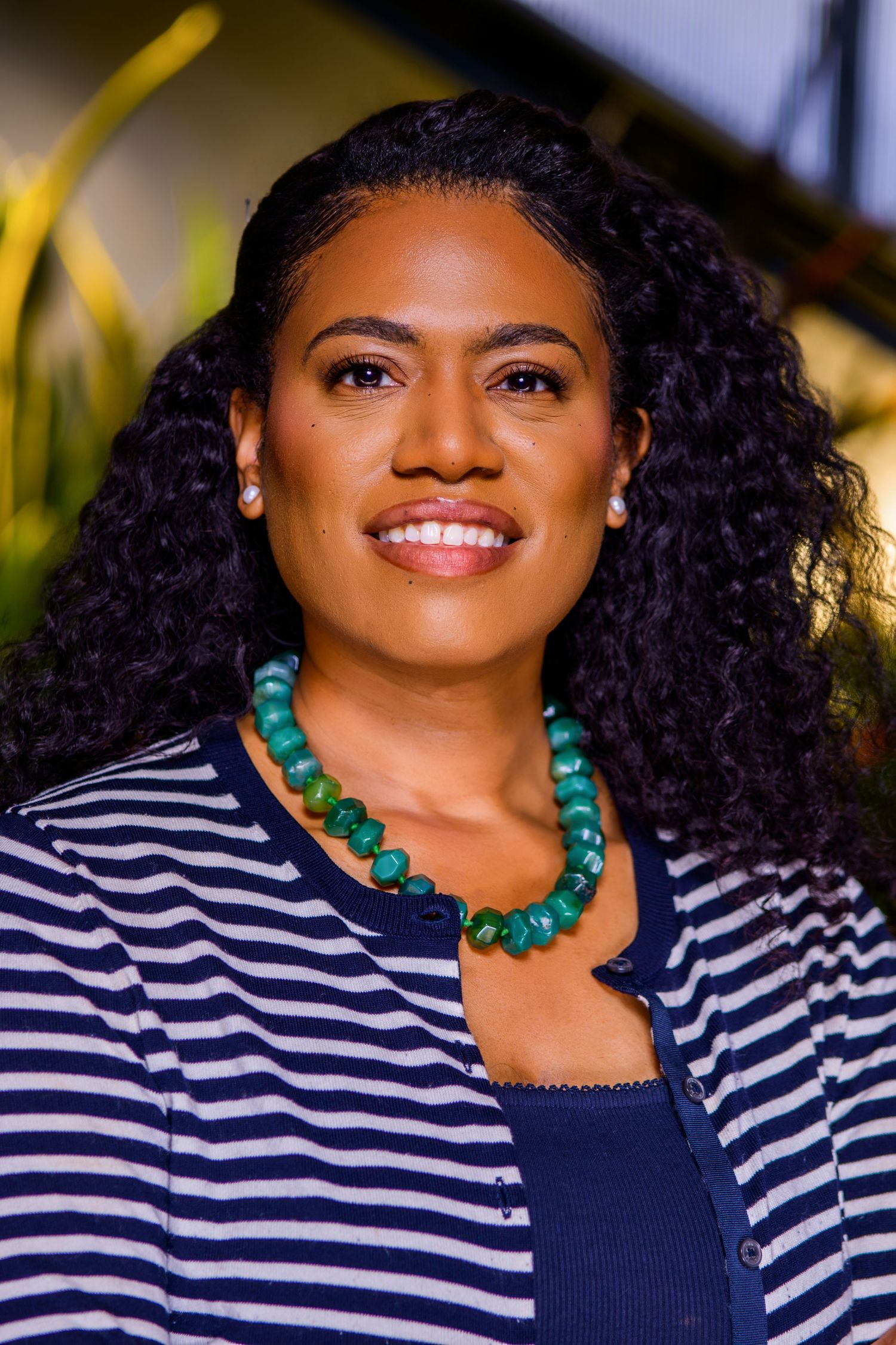 Smiling woman wearing a blue striped cardigan and green beaded necklace, with curly black hair, posing outdoors.