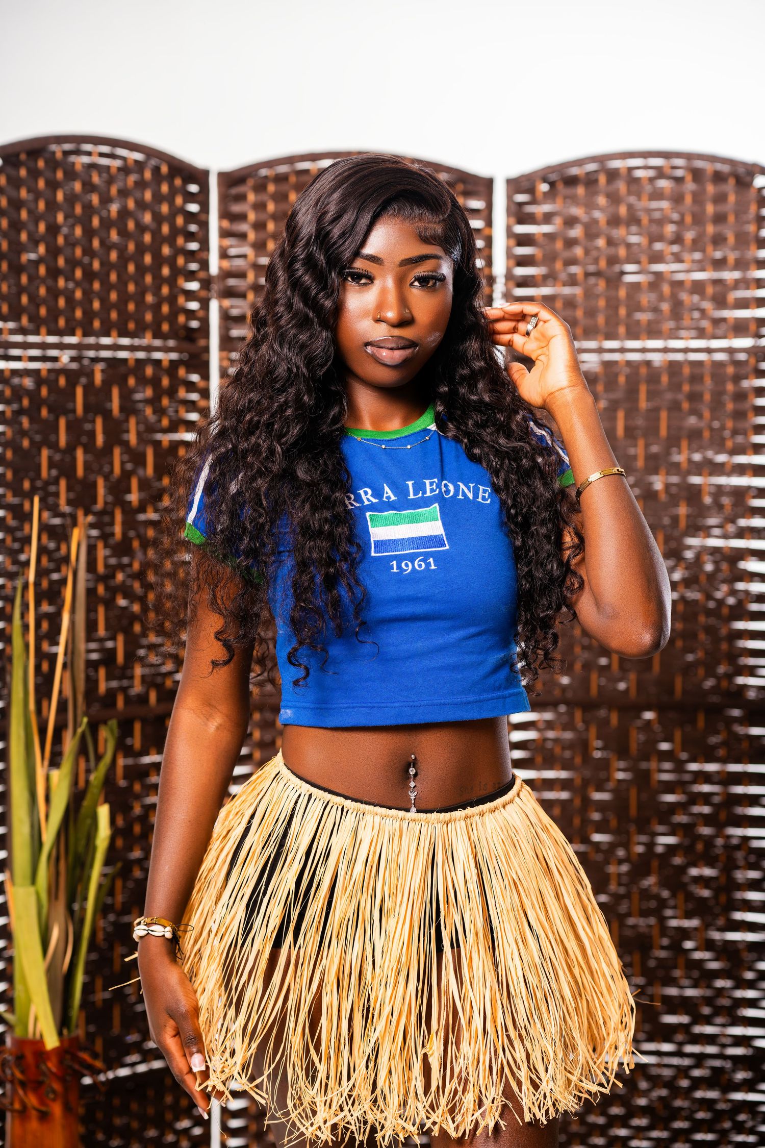 Young woman in Sierra Leone 1961 blue top and traditional grass skirt posing against woven brown backdrop.