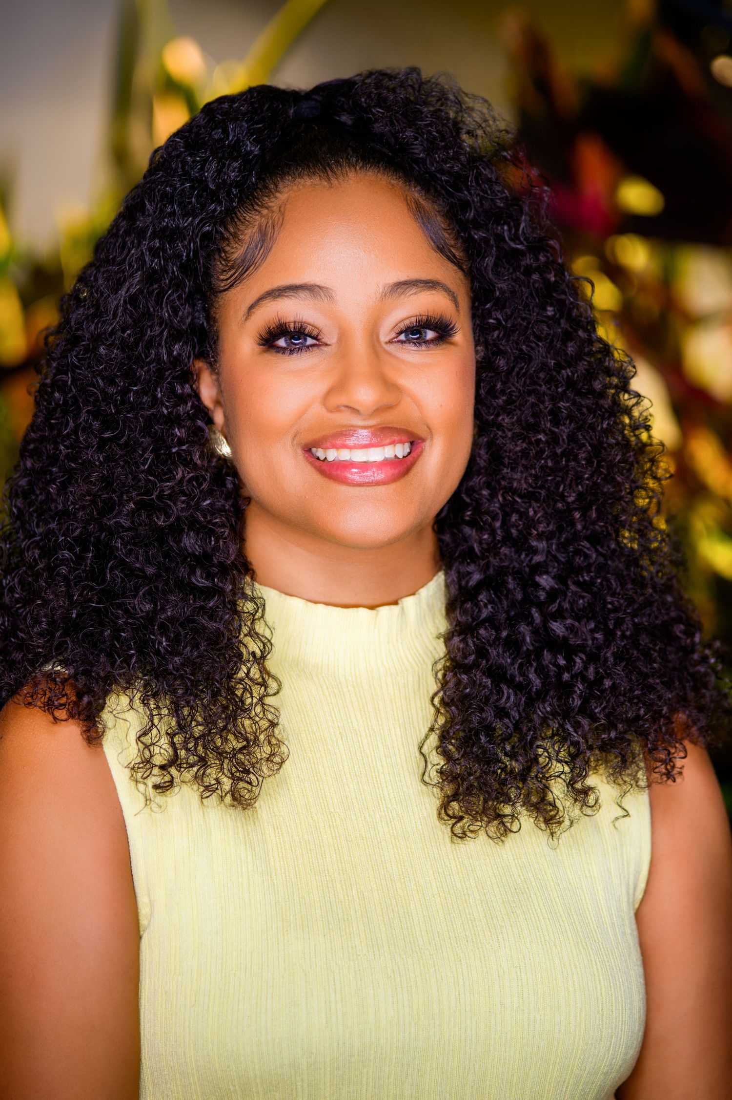 Smiling young woman with curly black hair wearing a light green sleeveless top, posed against a colorful floral background.