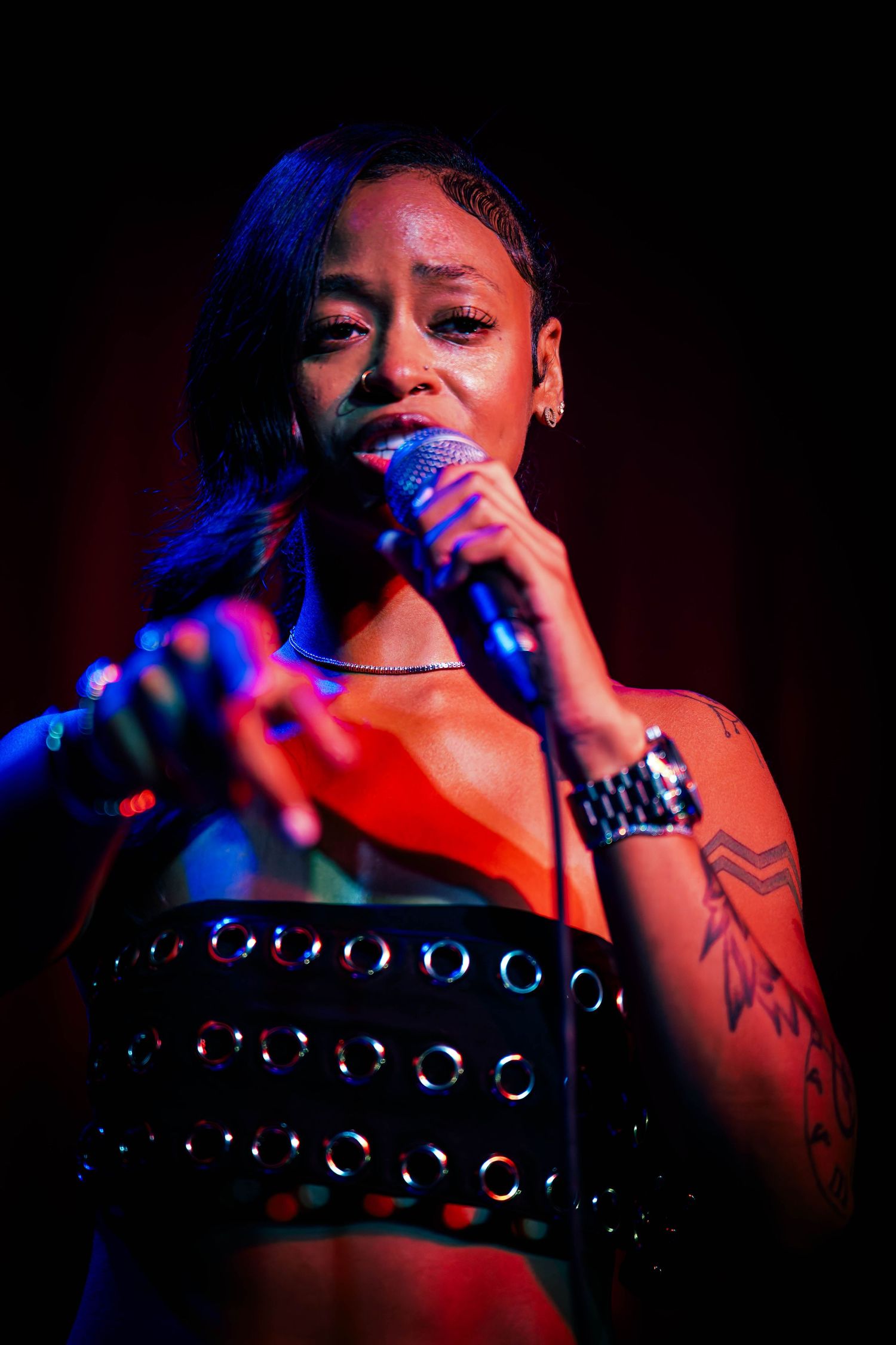 Singer performs on stage in dramatic lighting wearing a studded top with tattoos visible.