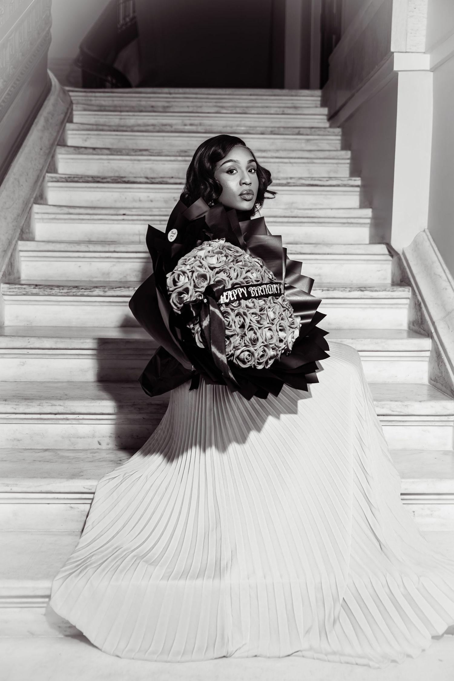 Young woman in pleated white skirt holding large floral bouquet on marble staircase, black and white photo.