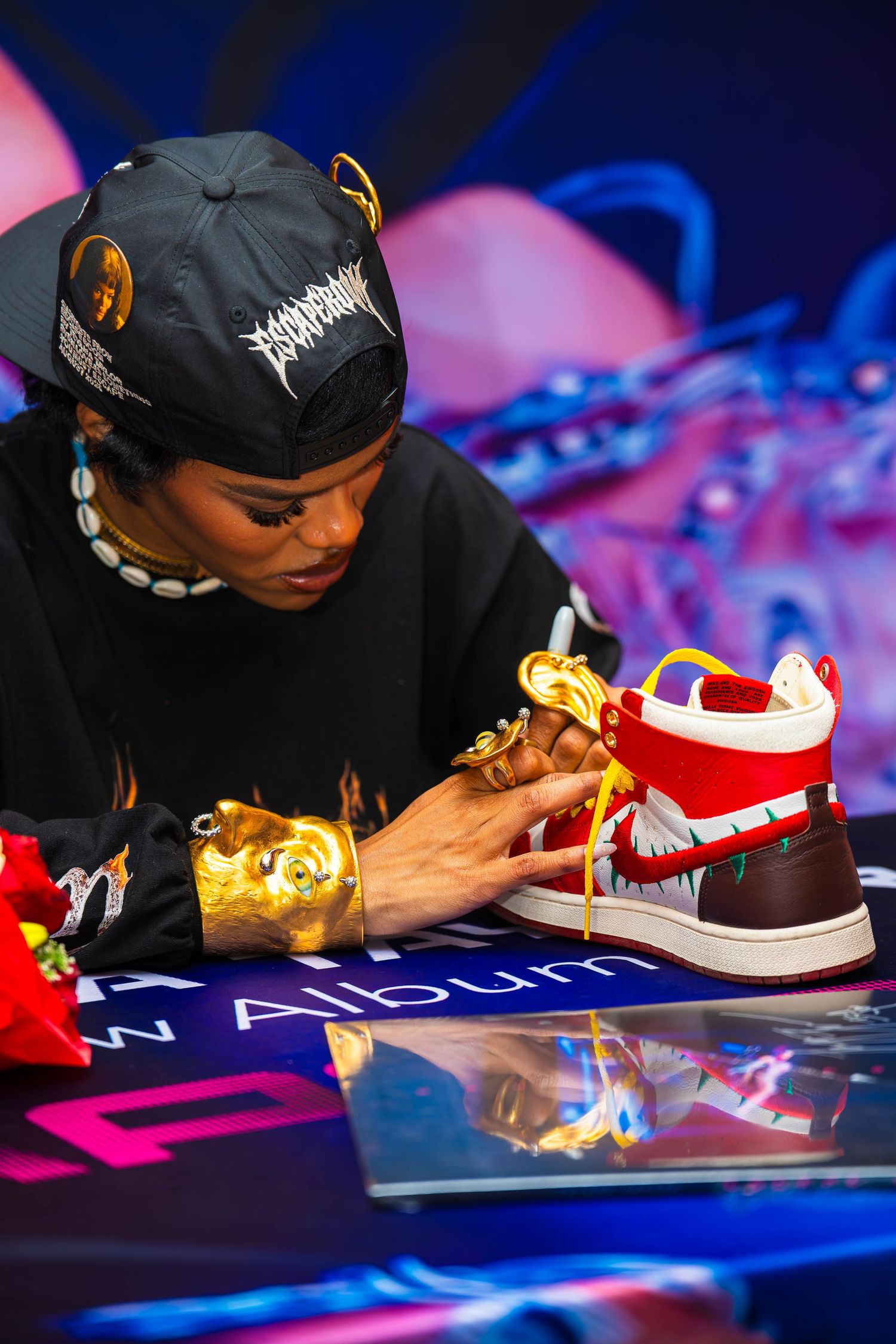Artist signing sneakers on table with colorful lighting and decorative elements.